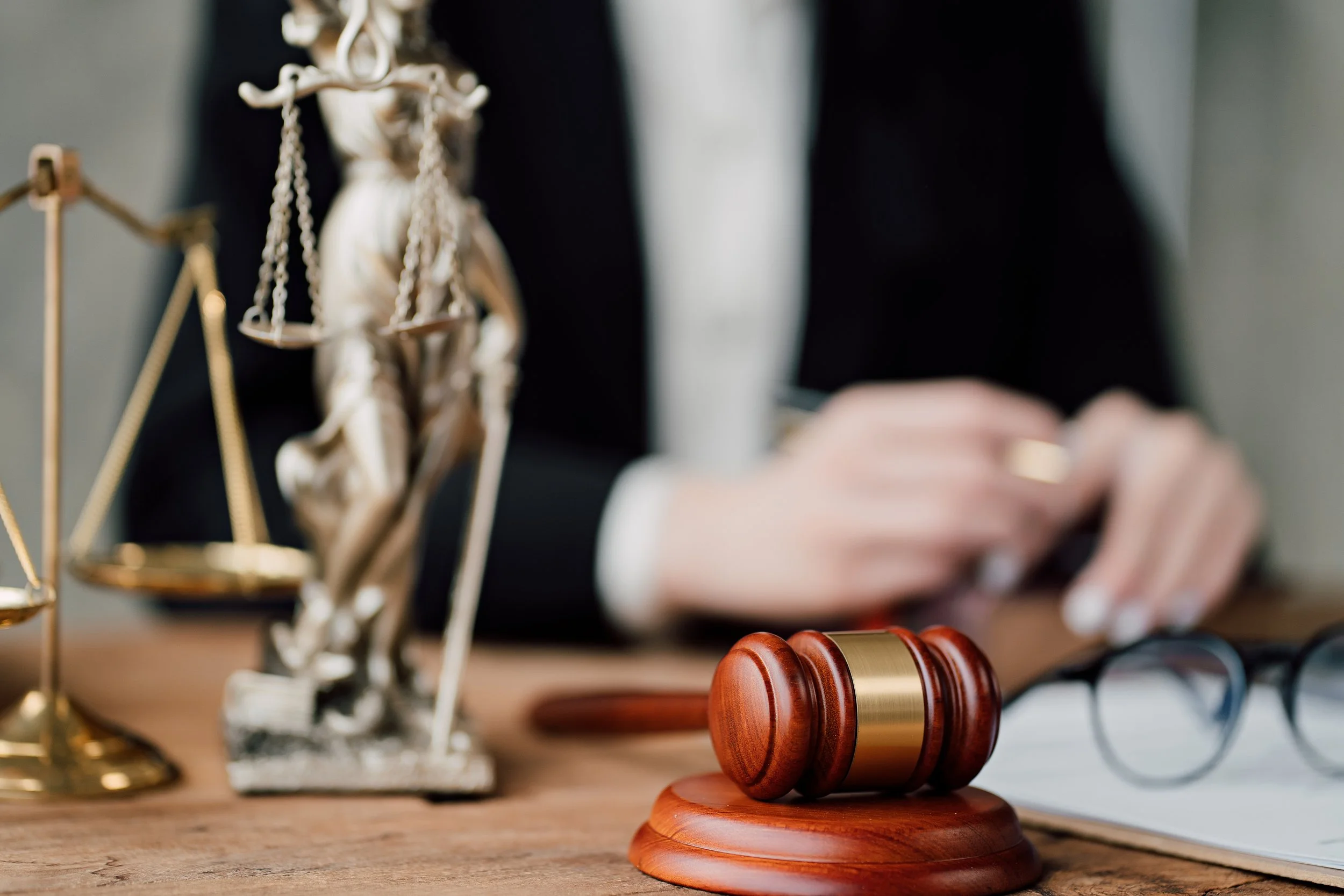 A judge's wooden gavel resting on a sound block on a wooden table, with a pair of eyeglasses and legal scales in the background, and a person in a suit blurred in the background.
