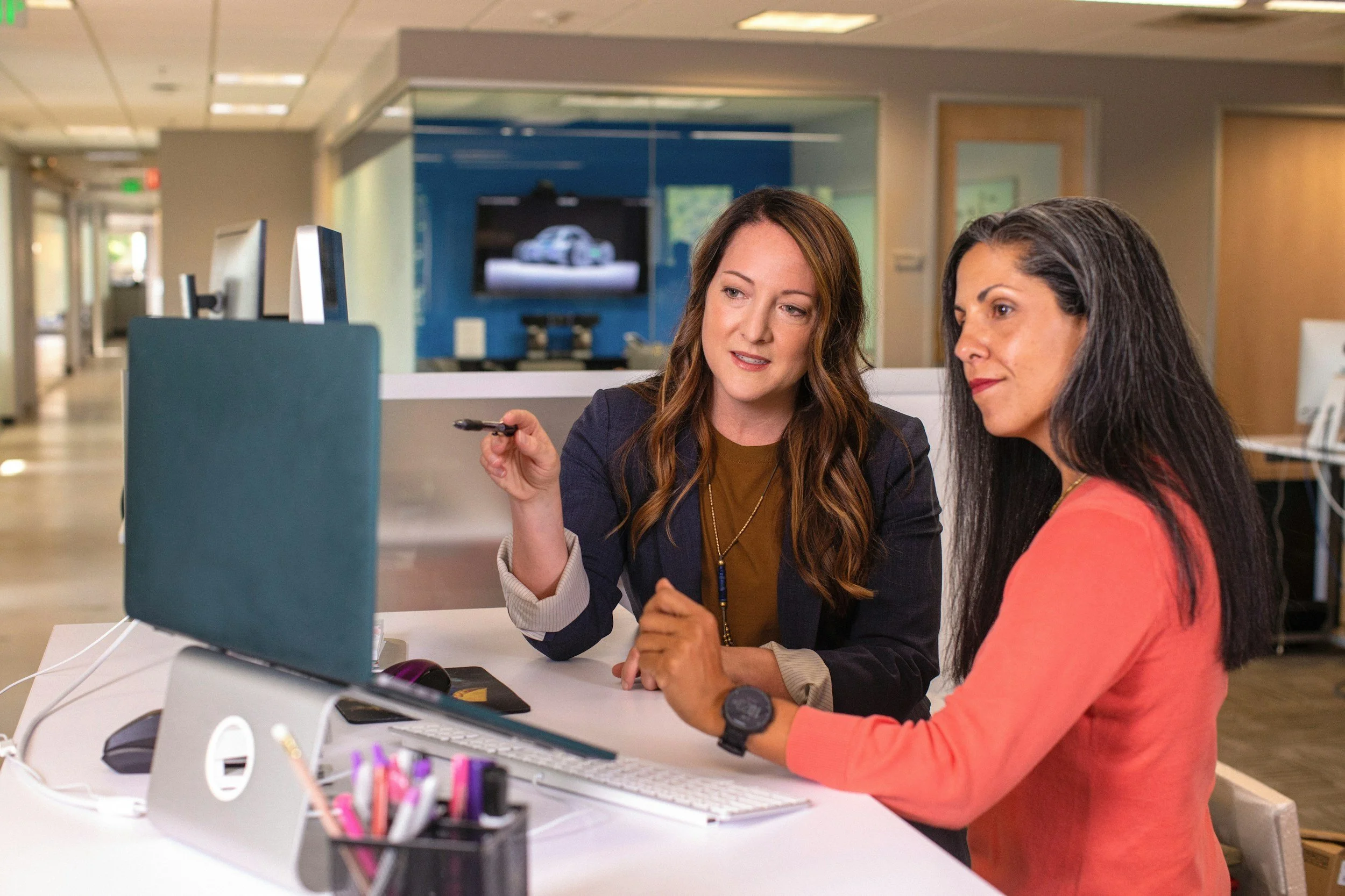 Two women sitting at an office desk, looking at a computer screen and discussing. One is holding a pen, and the other is wearing a smartwatch. The office has a modern setup with computers, office supplies, and background screens.