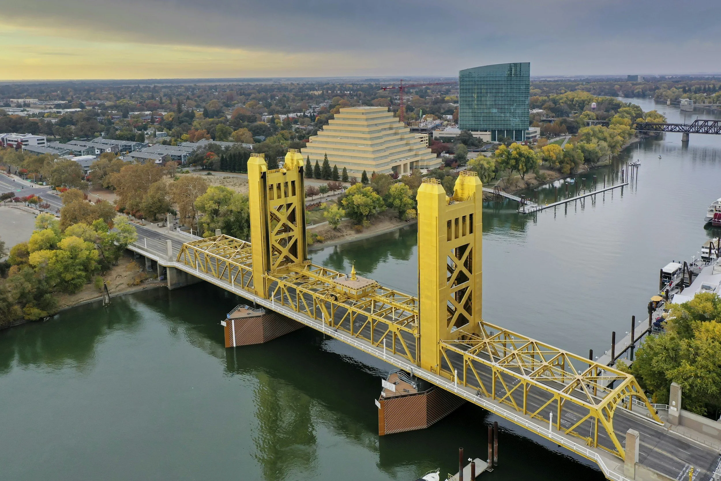 Aerial view of a yellow drawbridge crossing a river, with trees, modern buildings, and a stepped pyramid-like structure in the background.
