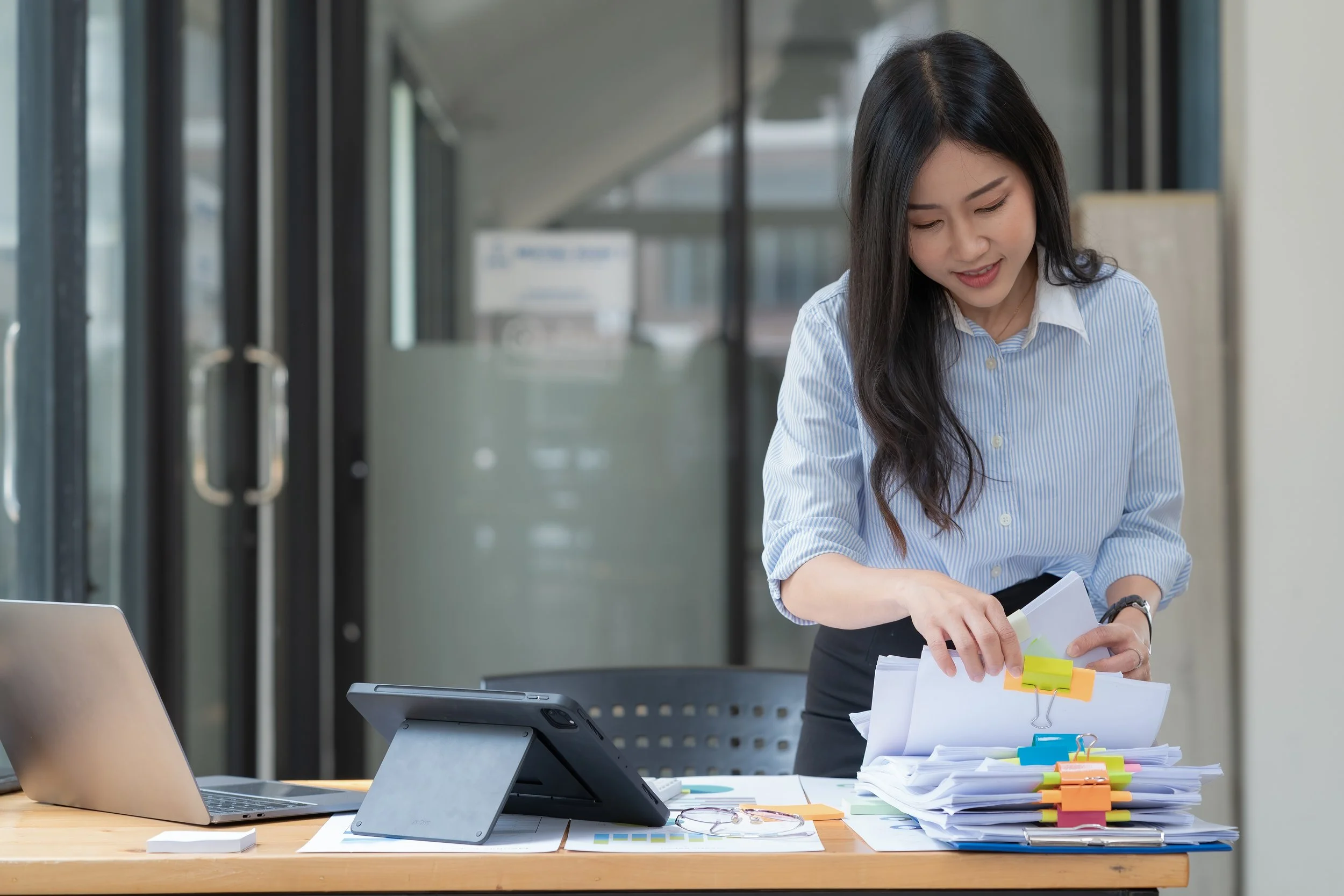A woman in business attire sorting through stacks of papers with colorful paper clips at a desk in an office, with a laptop and tablet on the desk.