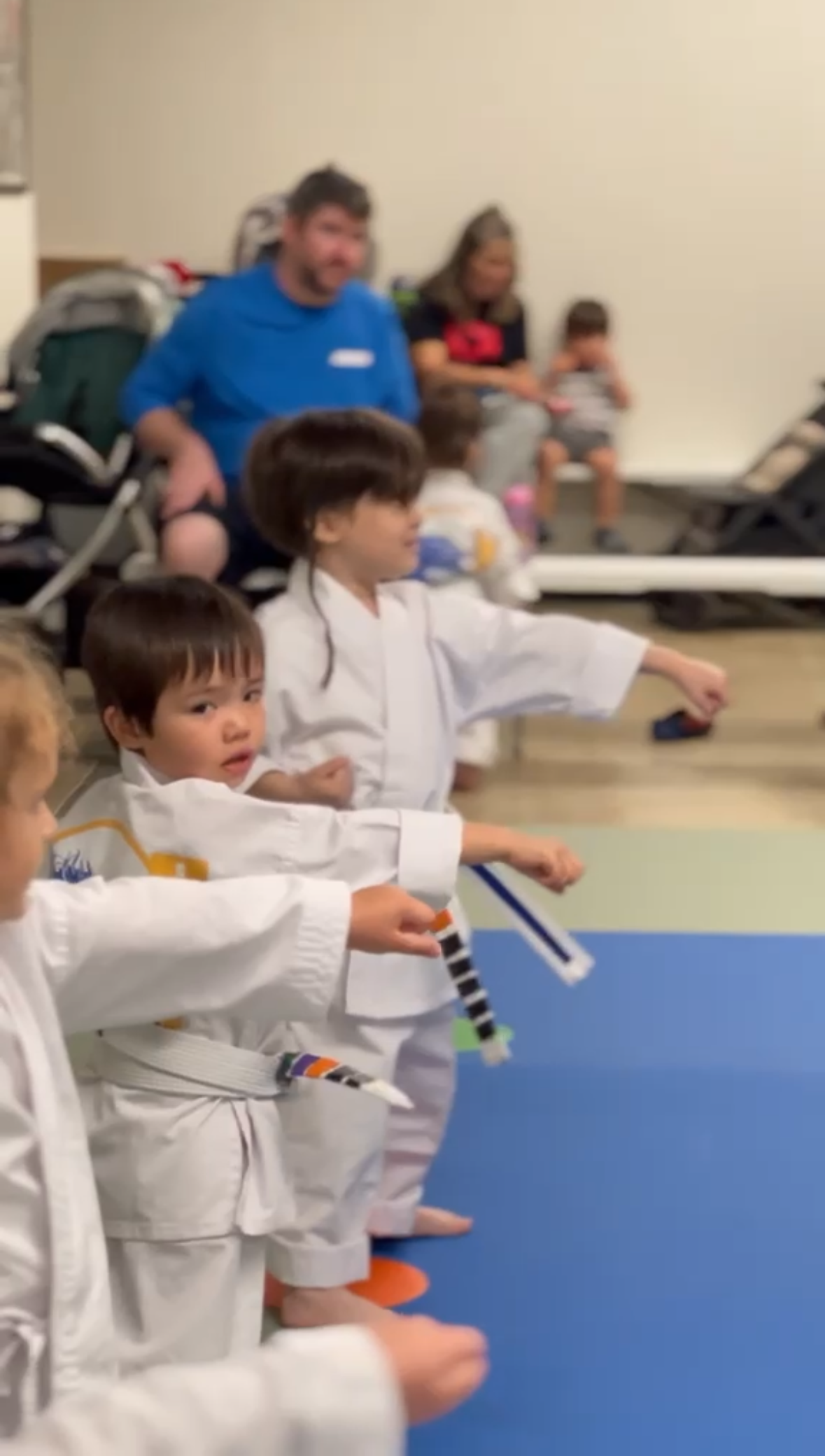 Children in martial arts uniforms practicing with striped sticks in a dojo, while adults watch in the background.