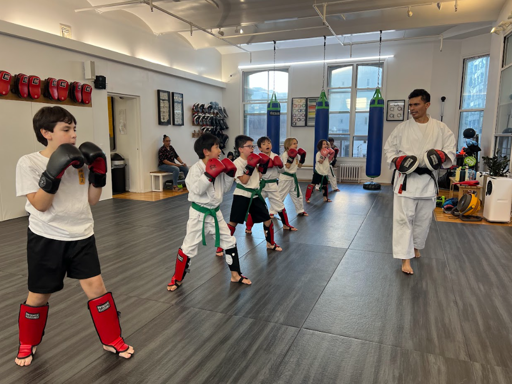 Martial arts class with children in white uniforms and green belts, practicing boxing with red and black gloves, led by an instructor in a white gi, in a gym with punching bags and equipment.