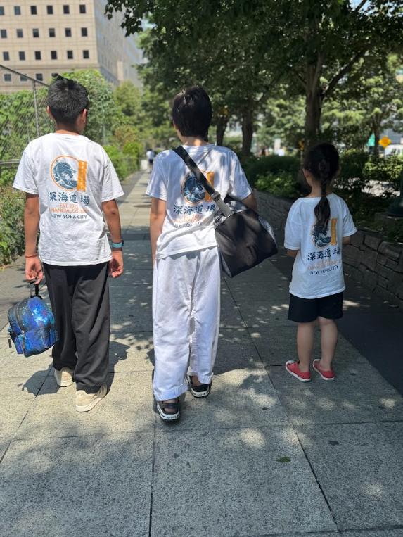 Three children, two girls and one boy, walking down a sidewalk in a city park, wearing white T-shirts with blue and orange graphics and text, carrying backpacks, with trees and tall buildings in the background.