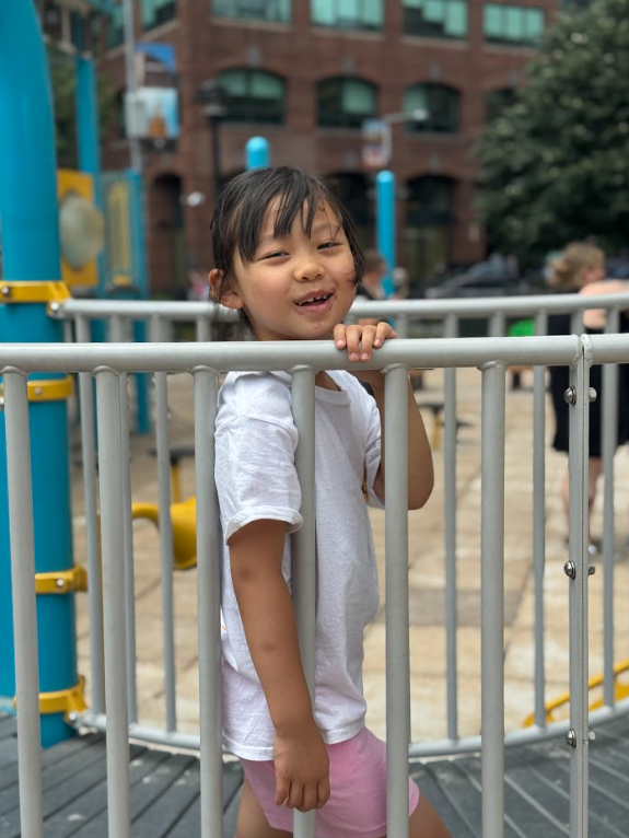 A young girl with dark hair wearing a white T-shirt and pink shorts standing behind a metal fence at a playground, smiling at the camera.