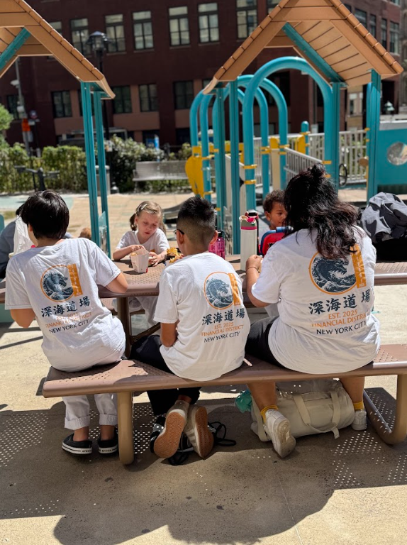 Four children and two adults sitting at a picnic table in a playground, with children eating and playing in the background.