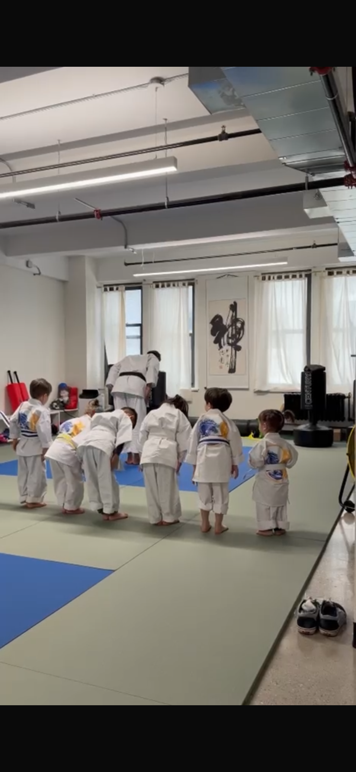 Children practicing martial arts in a dojo, with an instructor overseeing them, in a bright room with Japanese decor.