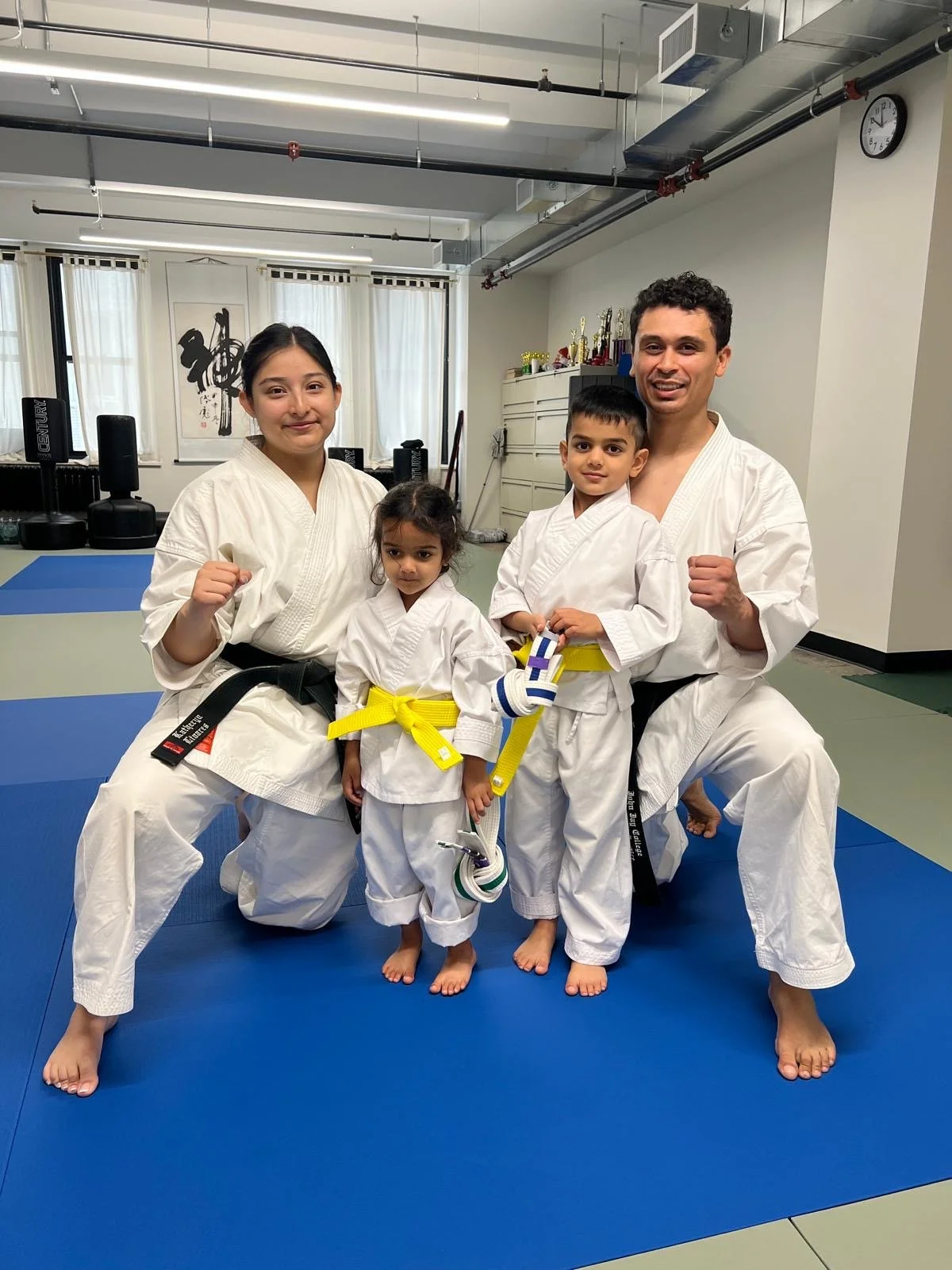 A group of four people in martial arts uniforms, kneeling and standing on a blue and green mat in a dojo, with trophies and calligraphy art on the wall in the background.