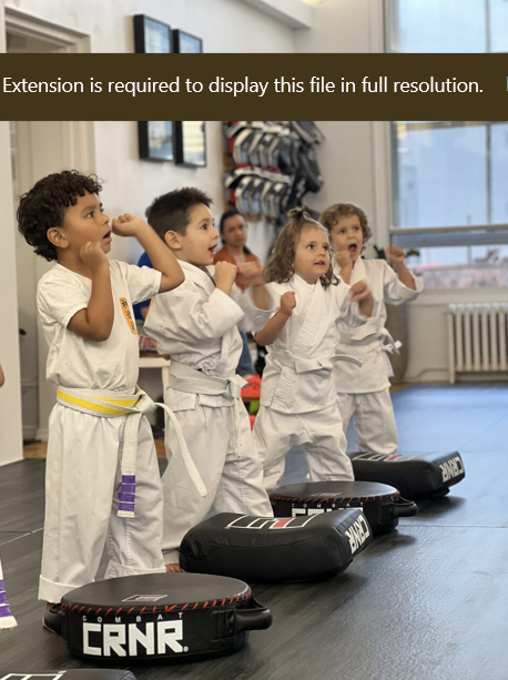 Children in martial arts uniforms (gi) practicing punches in a karate class, with focus pads in the foreground inside a dojo or gym.