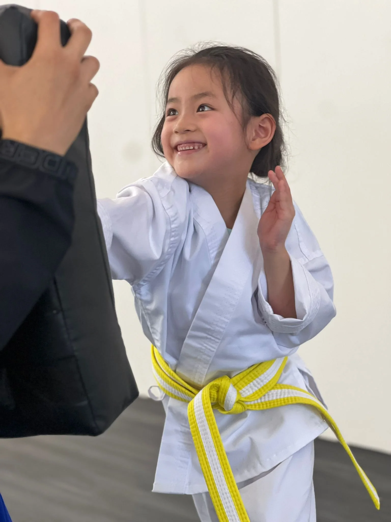 Young girl in a white martial arts gi and yellow belt smiling and posing for a photo in a martial arts dojo or training space.