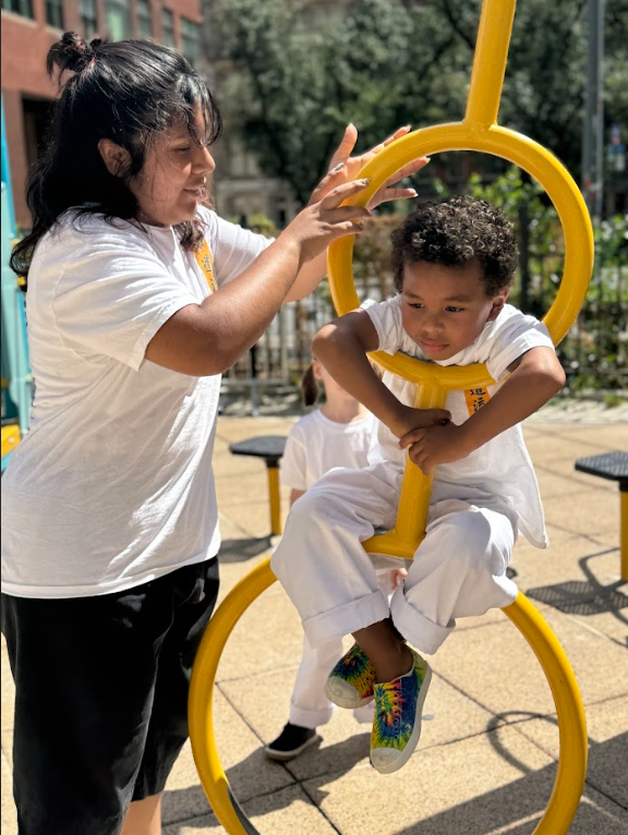 A young girl helping a boy sit on a yellow circular playground apparatus at an outdoor playground.