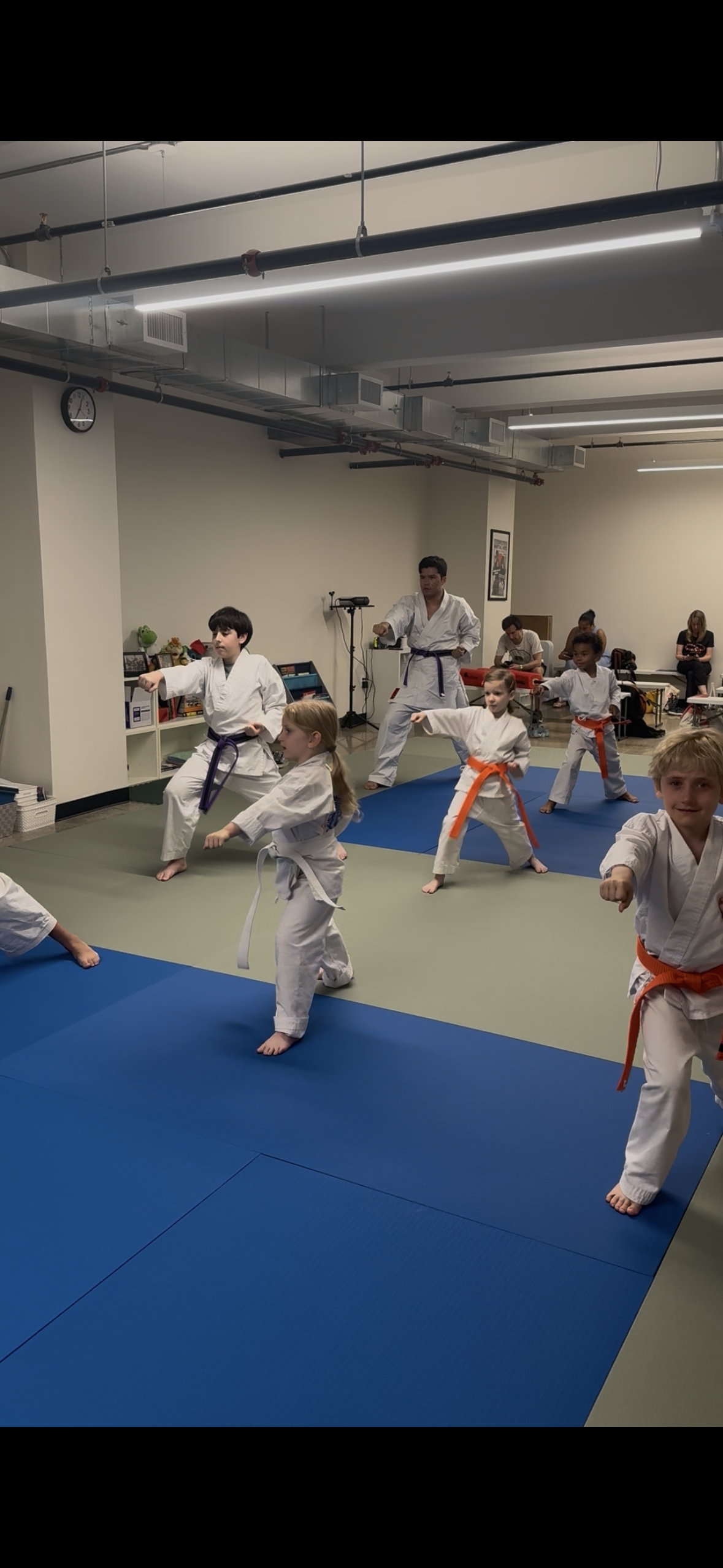 Children practicing karate in a dojo, supervised by a martial arts instructor, with adults sitting in the background.