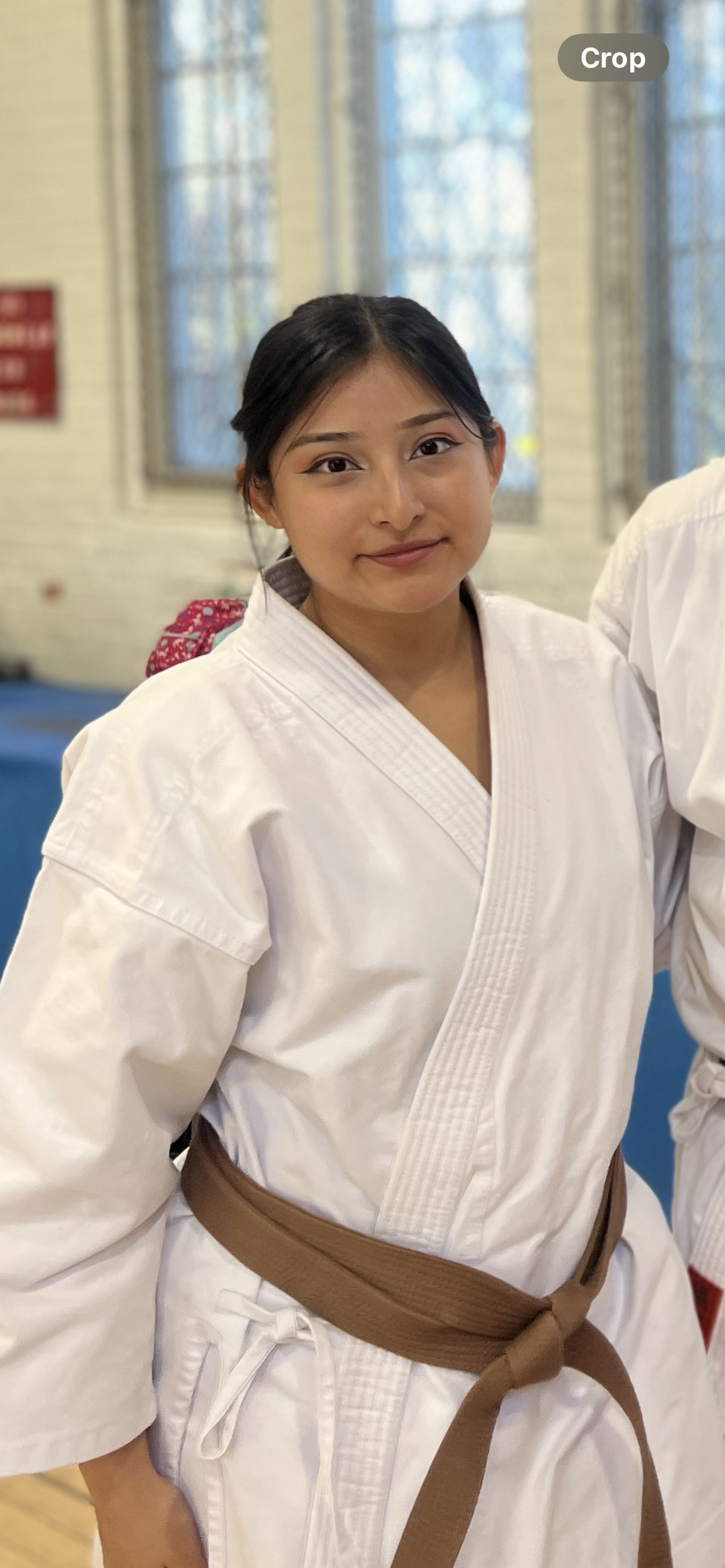 A young woman with dark hair styled in braids, wearing a white martial arts gi and a brown belt, stands in a dojo with a confident and happy expression.