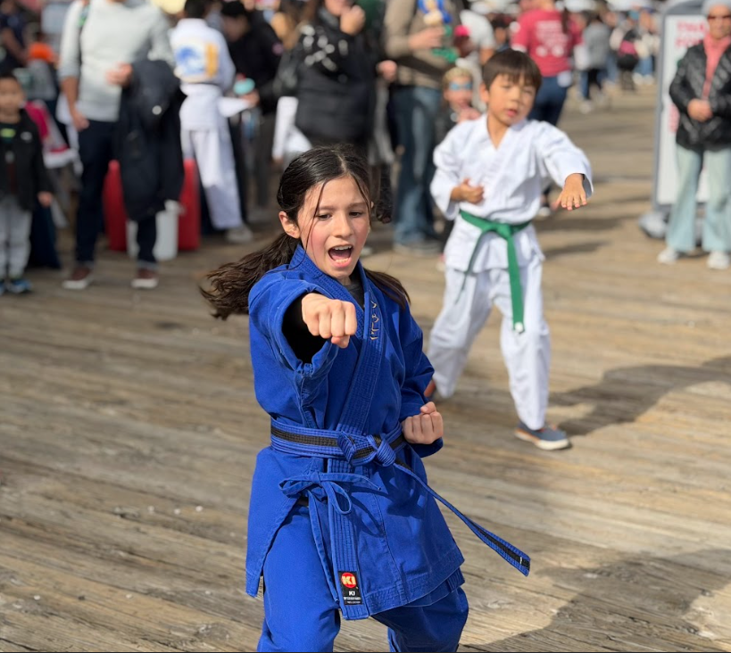 Young girl practicing karate wearing a blue gi on a wooden deck, punching forward with a fierce expression, with other children and spectators in the background.