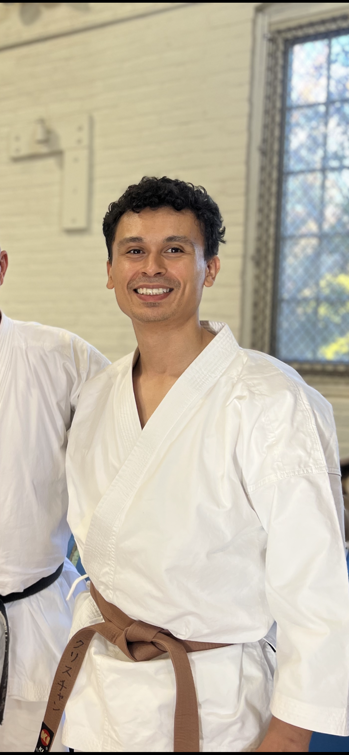 Person wearing a white martial arts gi with a brown belt, smiling indoors near a window.