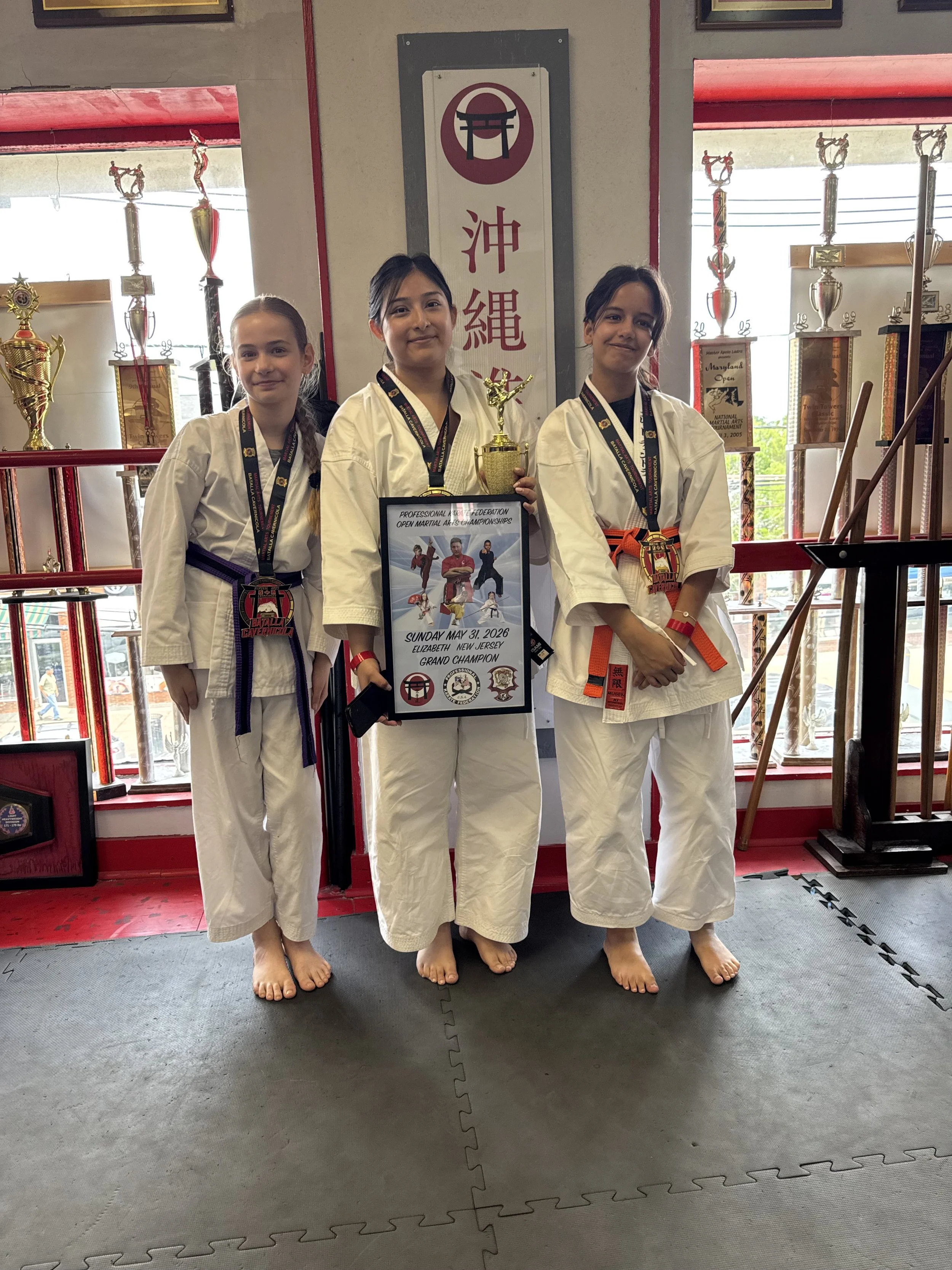 Three young girls in white karate uniforms with colored belts, standing side by side on martial arts mats inside a dojo, holding a trophy and a framed certificate, with numerous trophies displayed on shelves behind them.