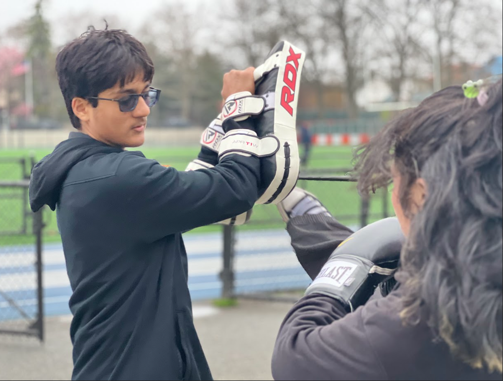 Young man in sunglasses helping another person with boxing gloves at an outdoor sports field.