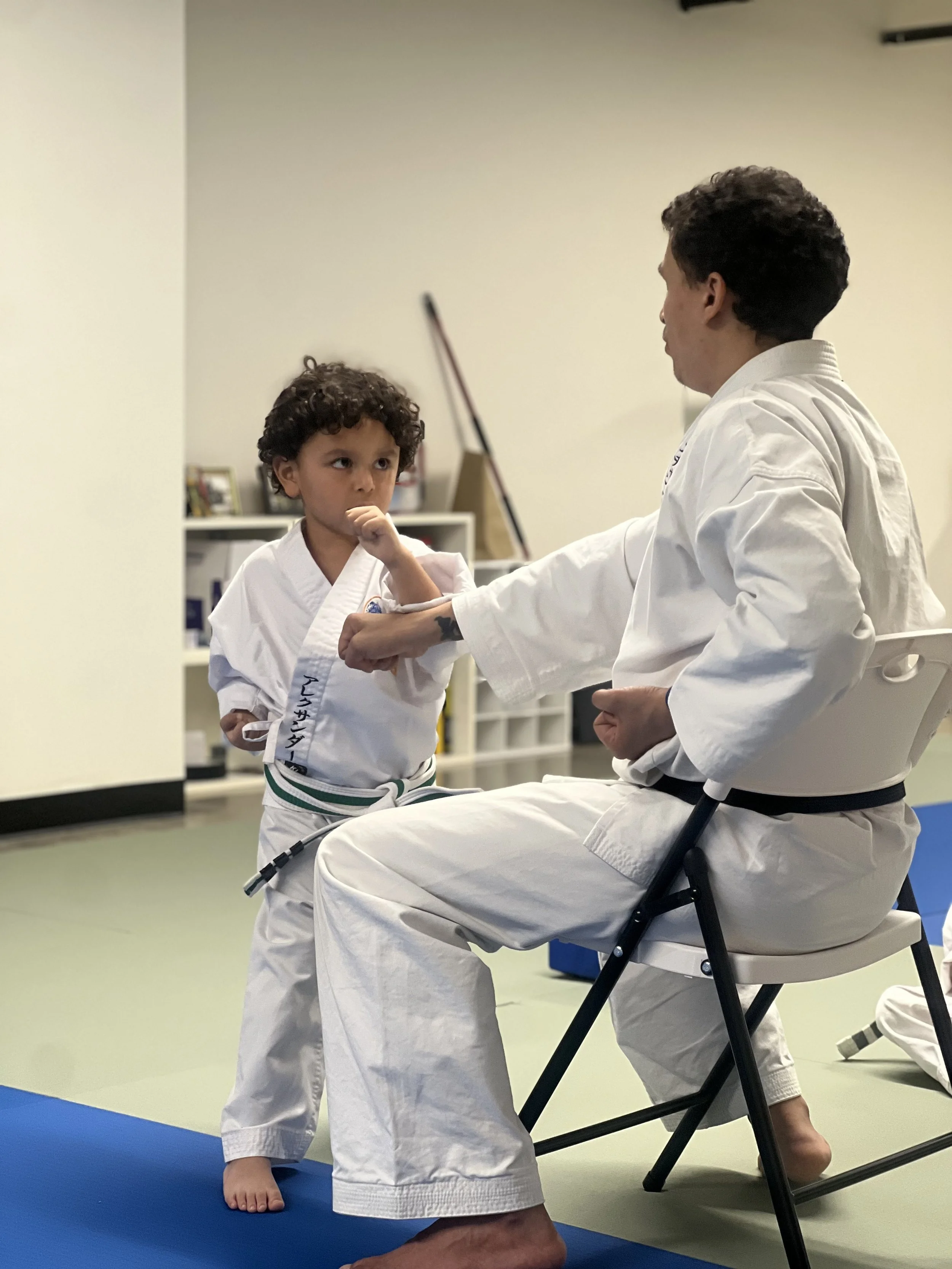 Two children practicing martial arts in a dojo, wearing white gi uniforms with green belts, with one sitting on a chair and the other standing.