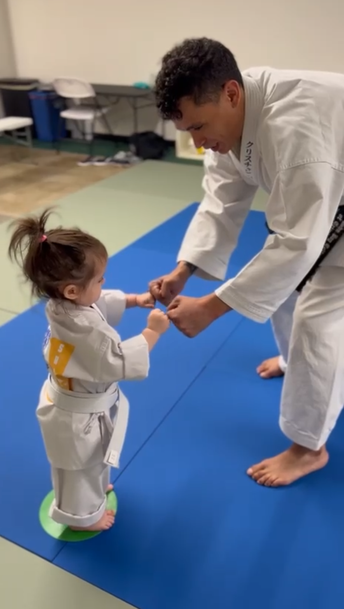 A young girl and an adult man practicing martial arts on a blue mat in a room. The girl is wearing a white martial arts uniform with a yellow belt, and the man is also in a white martial arts uniform. They are holding each other's fists in a training stance.