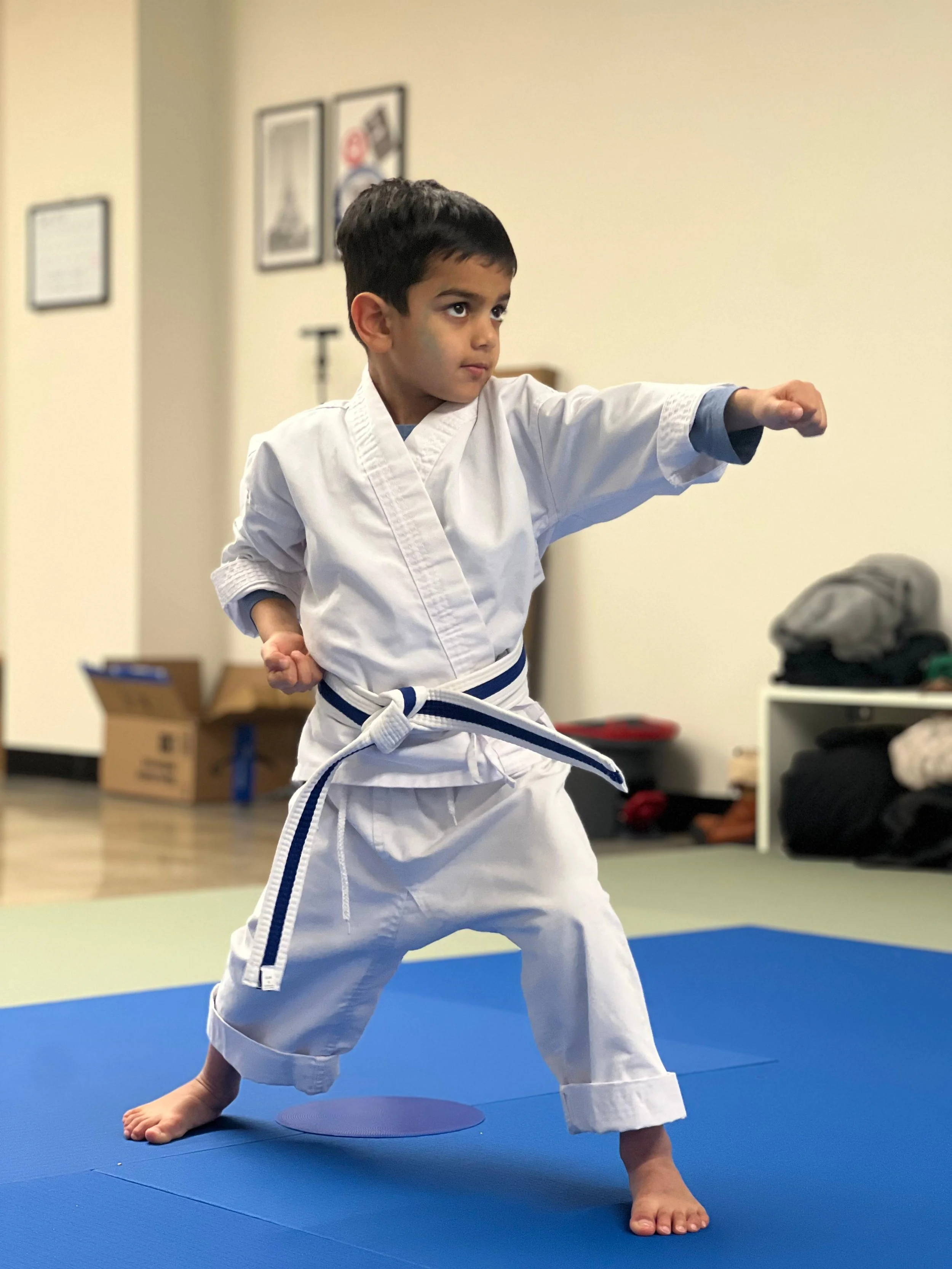 A young boy practicing martial arts in a white karate uniform with a blue belt, demonstrating a front punch stance on a blue mat in a dojo.