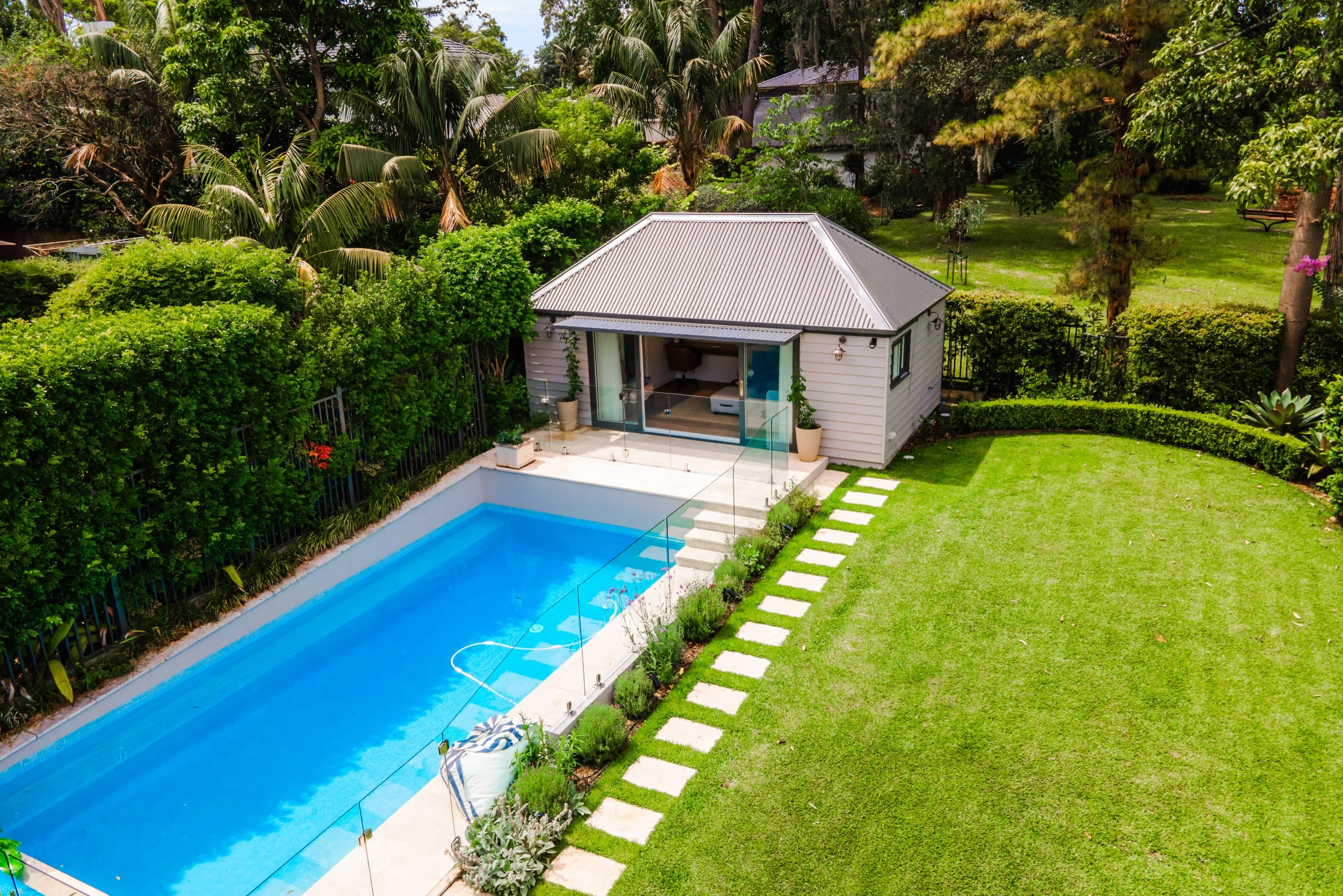 A backyard with a swimming pool, small house, and green lawn surrounded by trees and bushes.