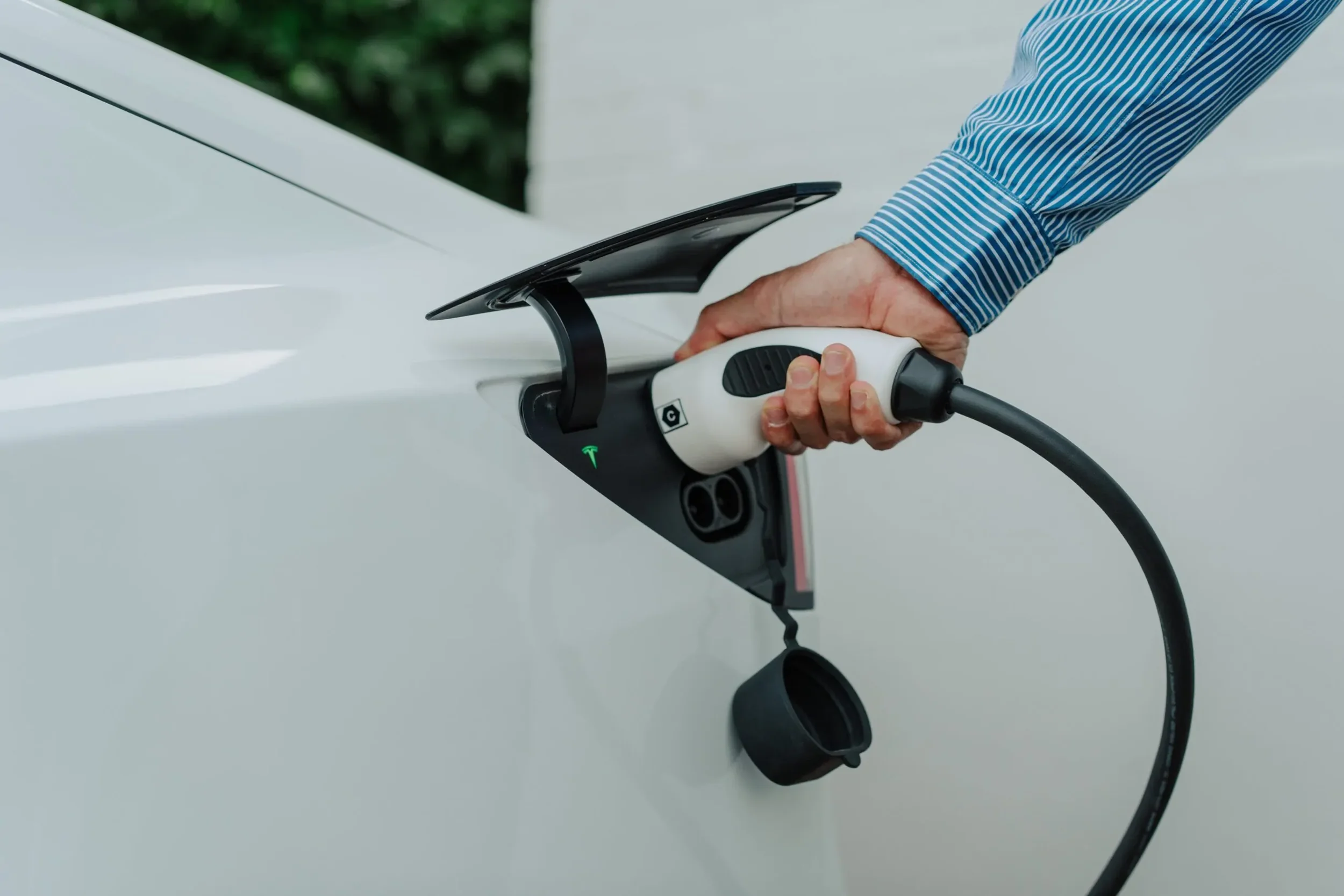 Person charging a white Tesla electric vehicle using a charging station.