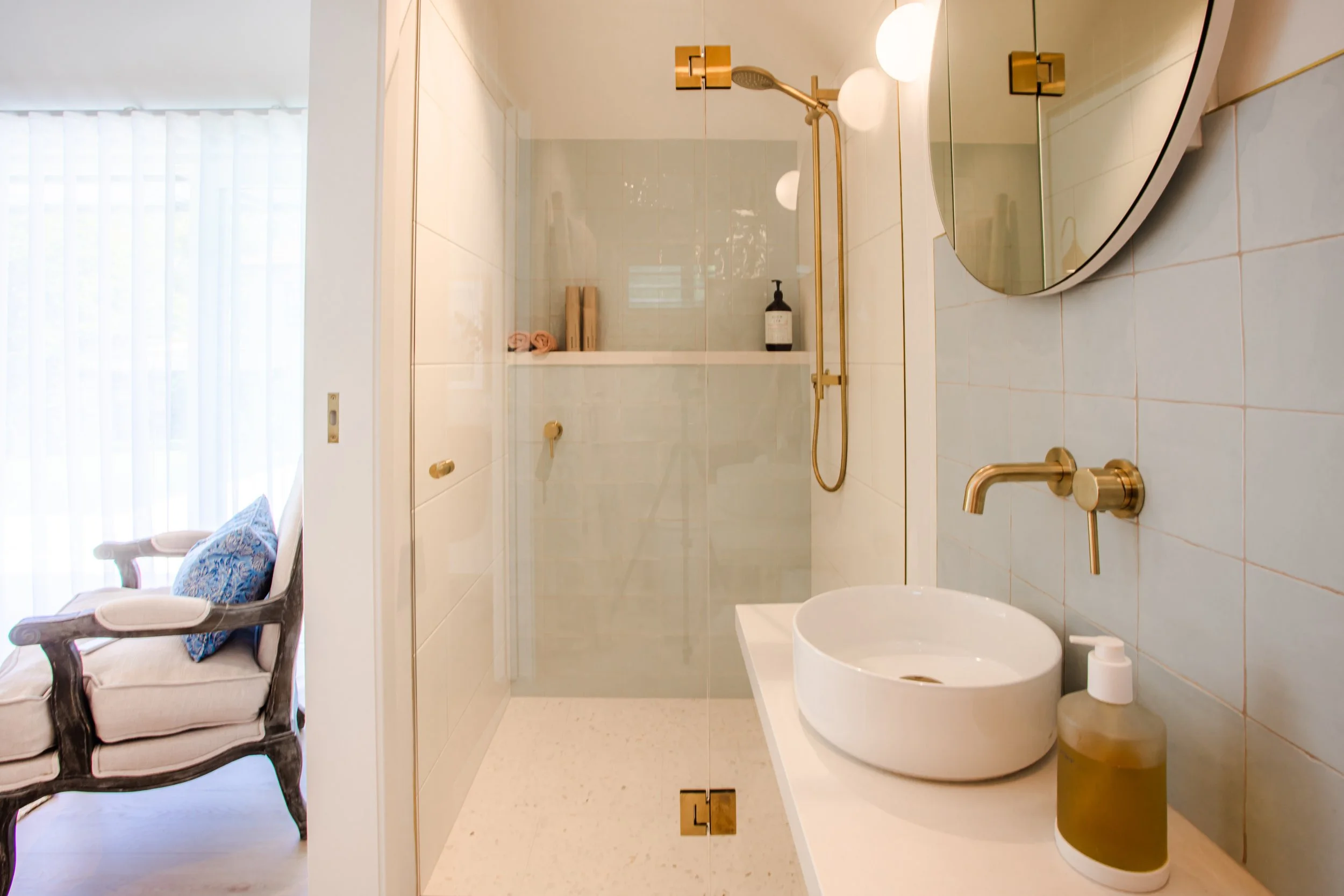 Modern bathroom with a round mirror, wall-mounted brass faucet, white vessel sink, soap dispenser, and a glass shower enclosure with brass fixtures, next to a sitting area with a vintage armchair and cushion.