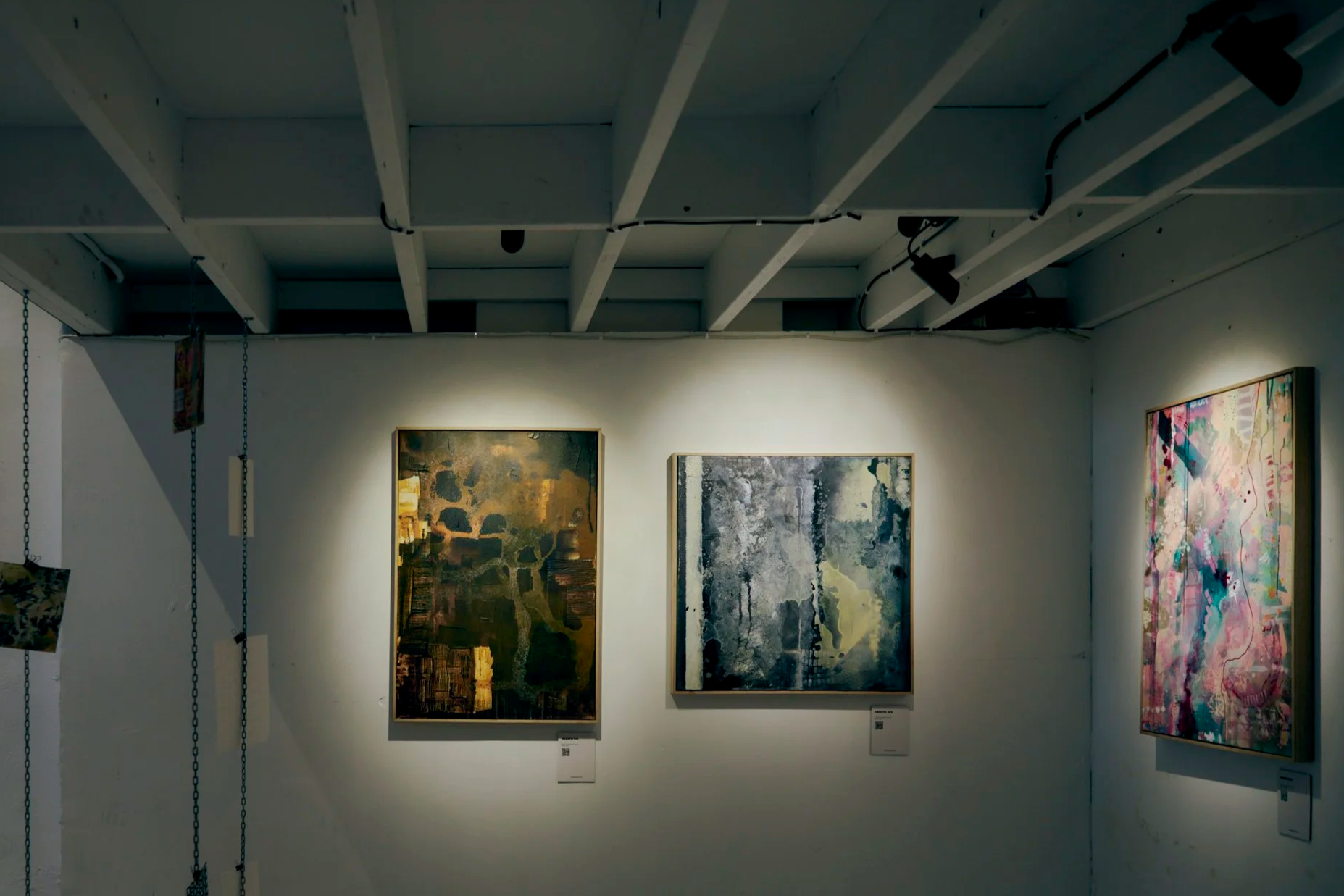 Three abstract paintings hanging on a white gallery wall illuminated by overhead lighting in an art exhibition.