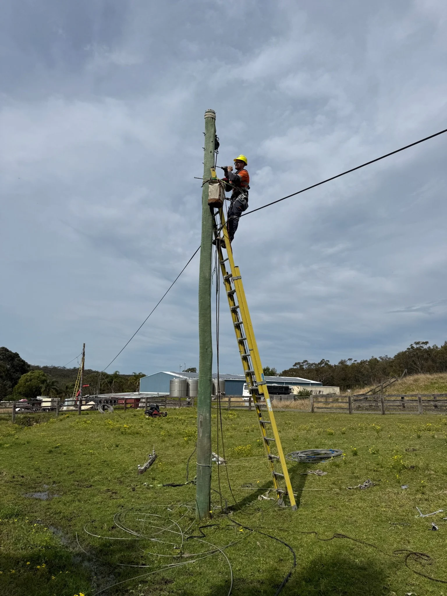A worker in safety gear, including a yellow helmet, is standing on a ladder fixing a utility pole in a grassy field with a cloudy sky in the background.