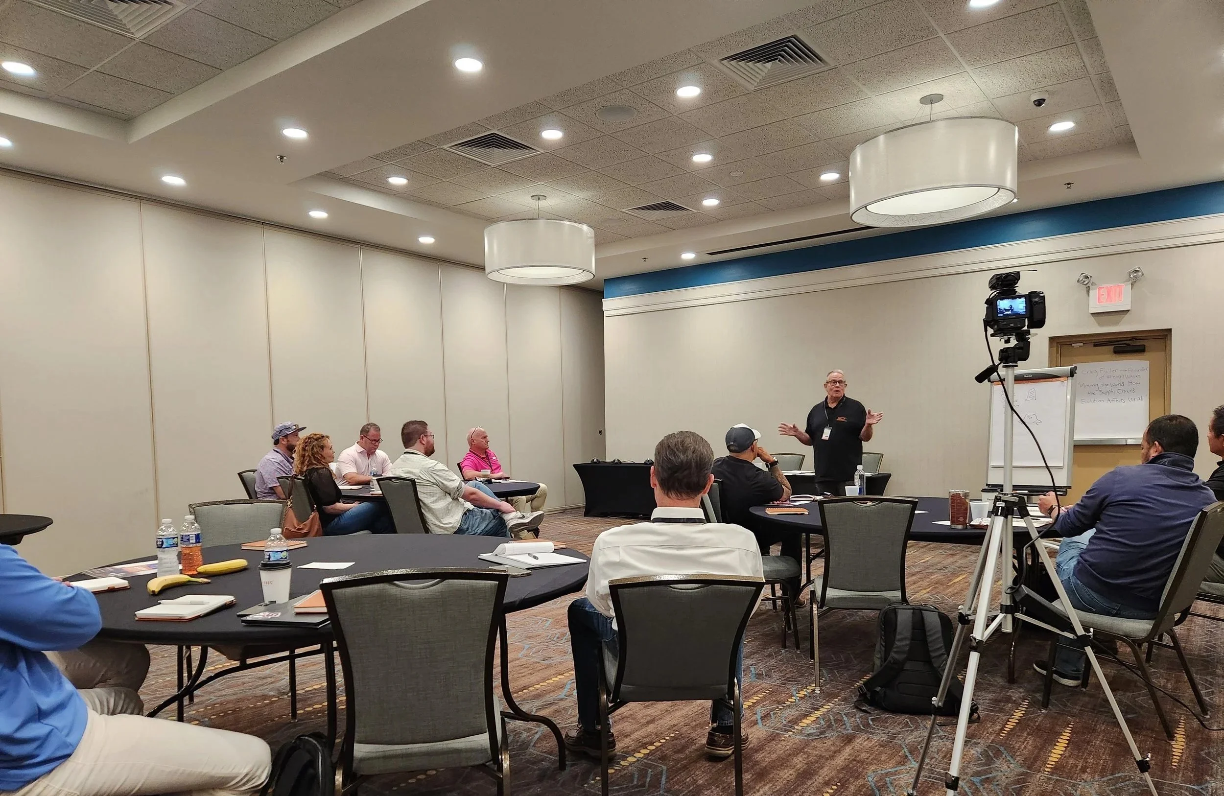 A man is standing in front of a small group in a conference room, giving a presentation. Attendees are seated at round tables with notebooks, water bottles, and bananas. A camera on a tripod records the event.