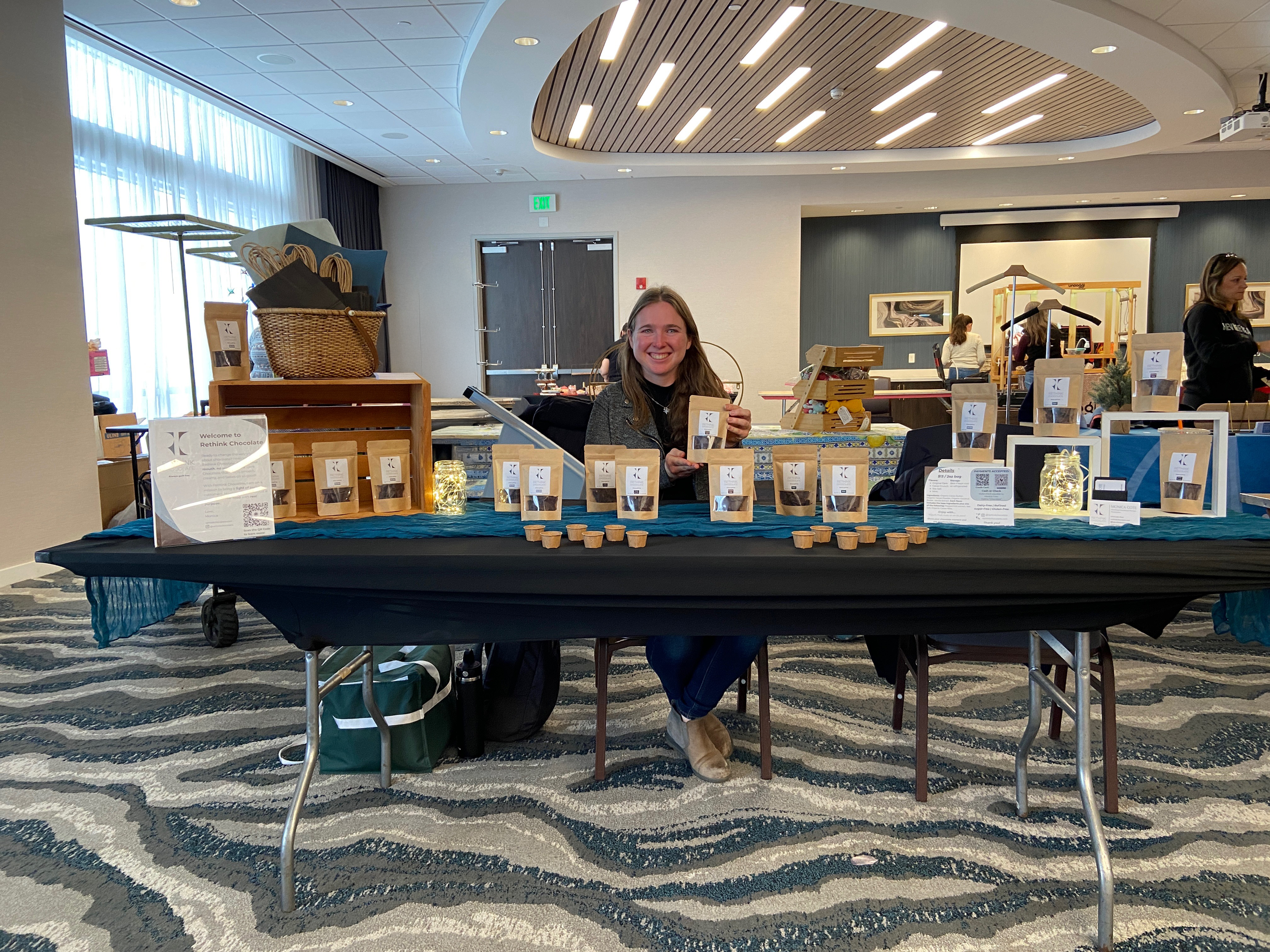 Woman sitting at a table with various packages of chocolate, smiling and holding a piece of chocolate, in a conference or event space.
