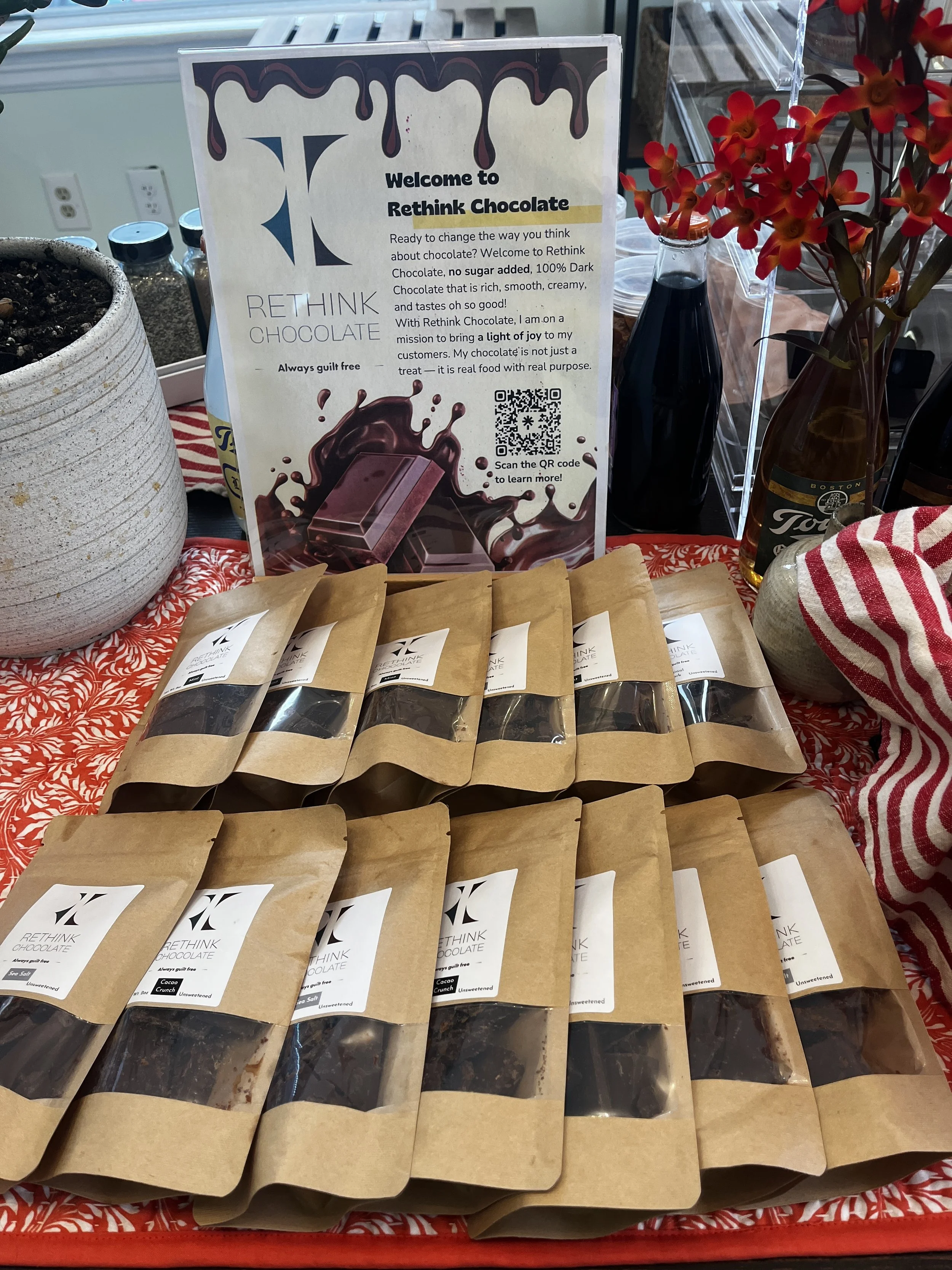 Display of various bags of Rethink Chocolate on a red patterned tablecloth, with a sign explaining the product and a QR code. Decorative flowers and bottles are in the background.