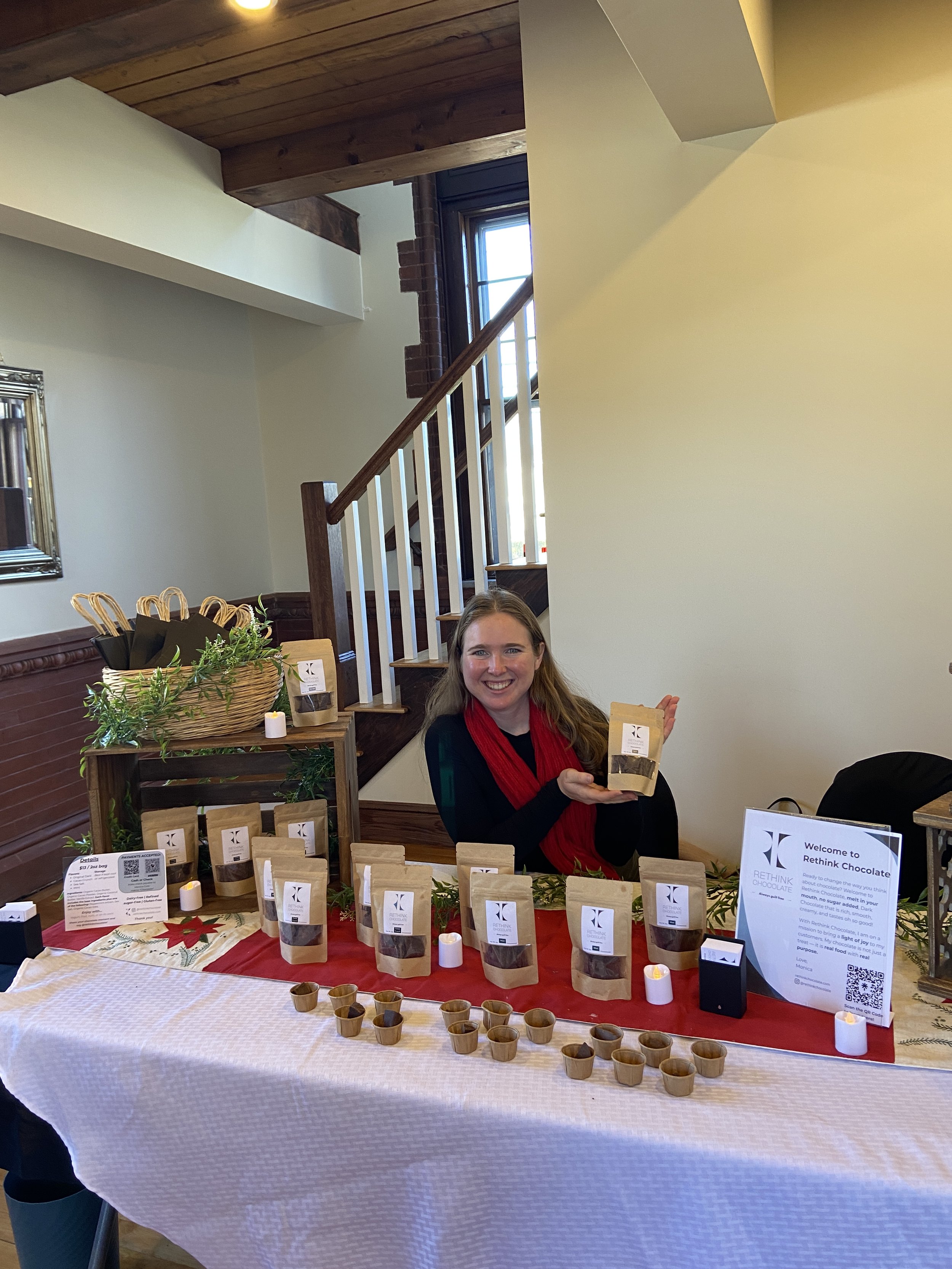 A woman with long hair and a red scarf smiling while sitting at a table displaying chocolate products for sale. The table has a red tablecloth, small tasting cups, and a sign welcoming customers to Rethink Chocolate. The background includes a staircase and a wall with a mirror.
