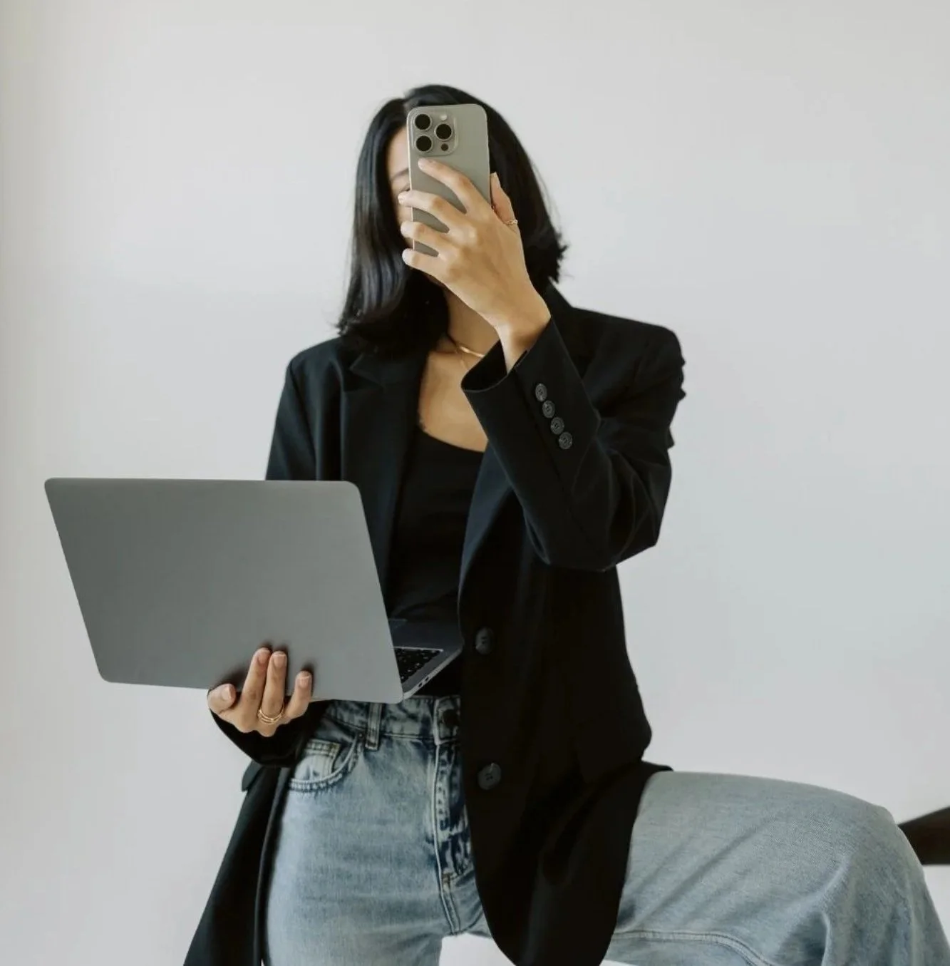 Person in a black blazer holding a laptop in one hand and taking a photo with a smartphone in front of a plain white background.