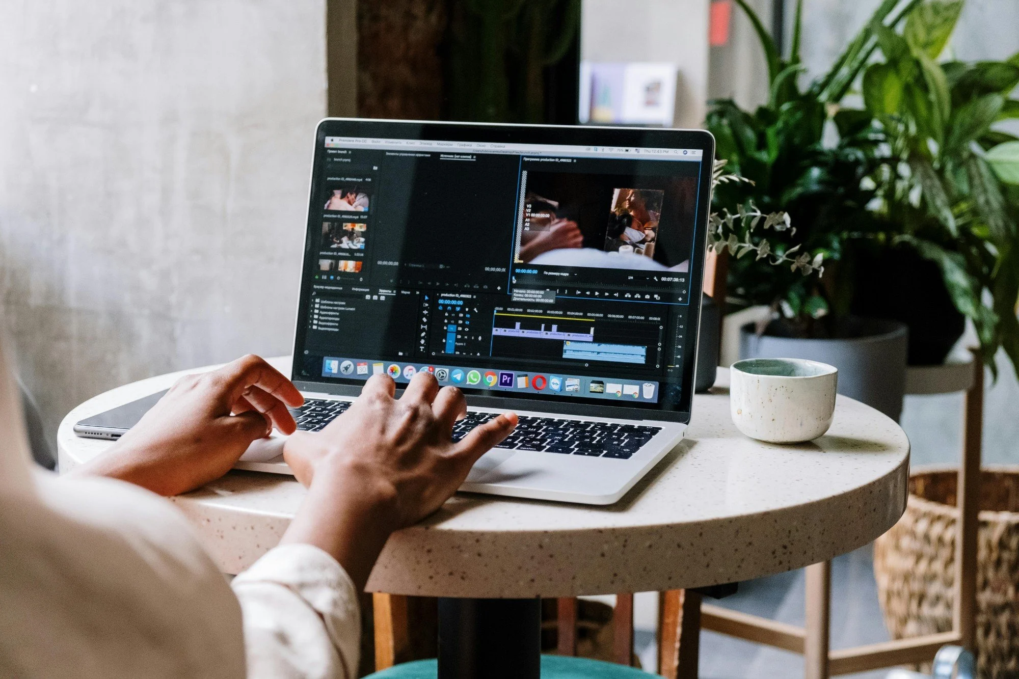 Person working on a video editing project on a MacBook laptop at a small round table, with a coffee cup beside the laptop and green plants in the background.