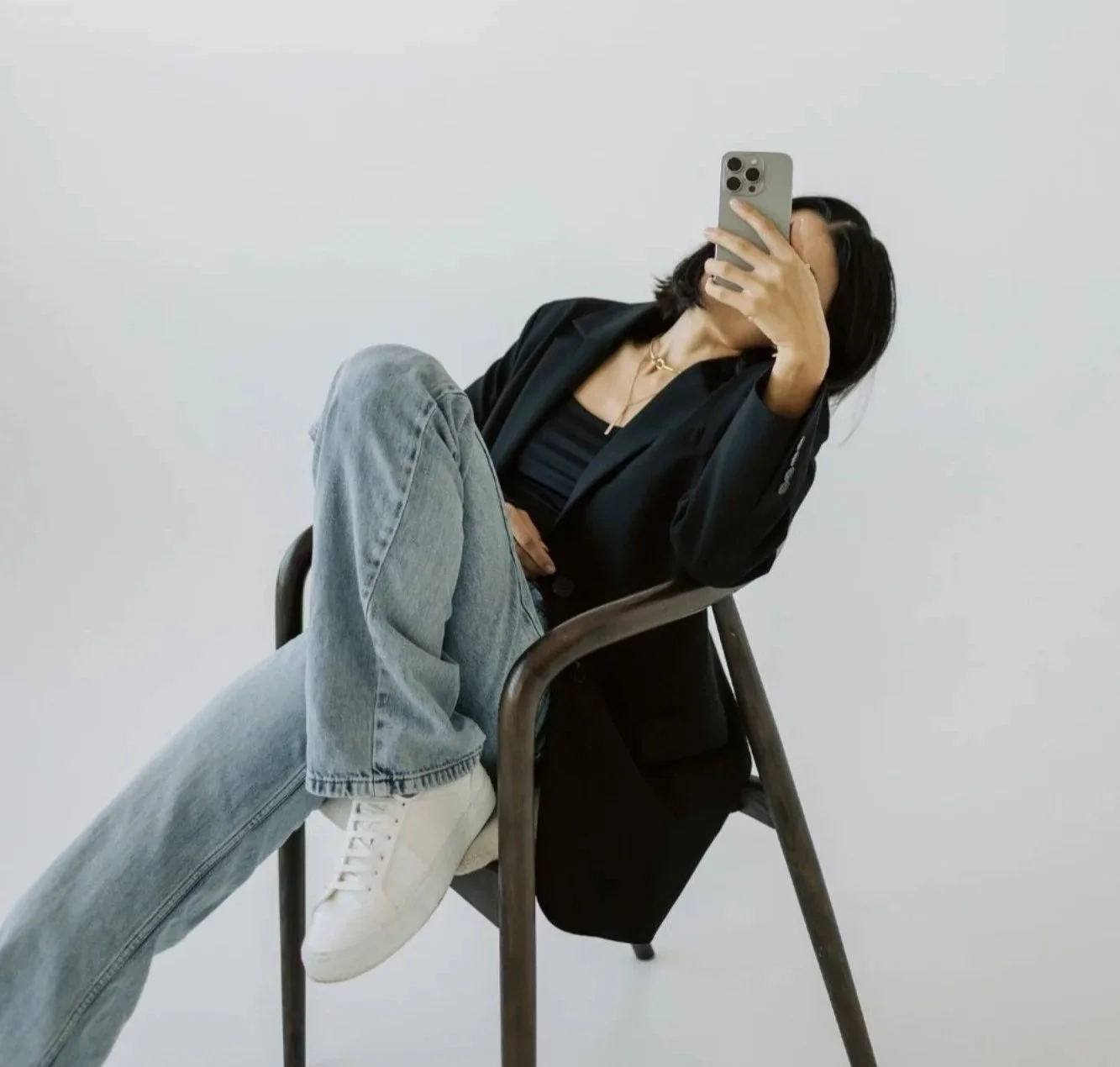 Woman sitting on a chair, taking a selfie with her phone, wearing a black blazer, black top, light-wash jeans, and white sneakers, against a plain white background.