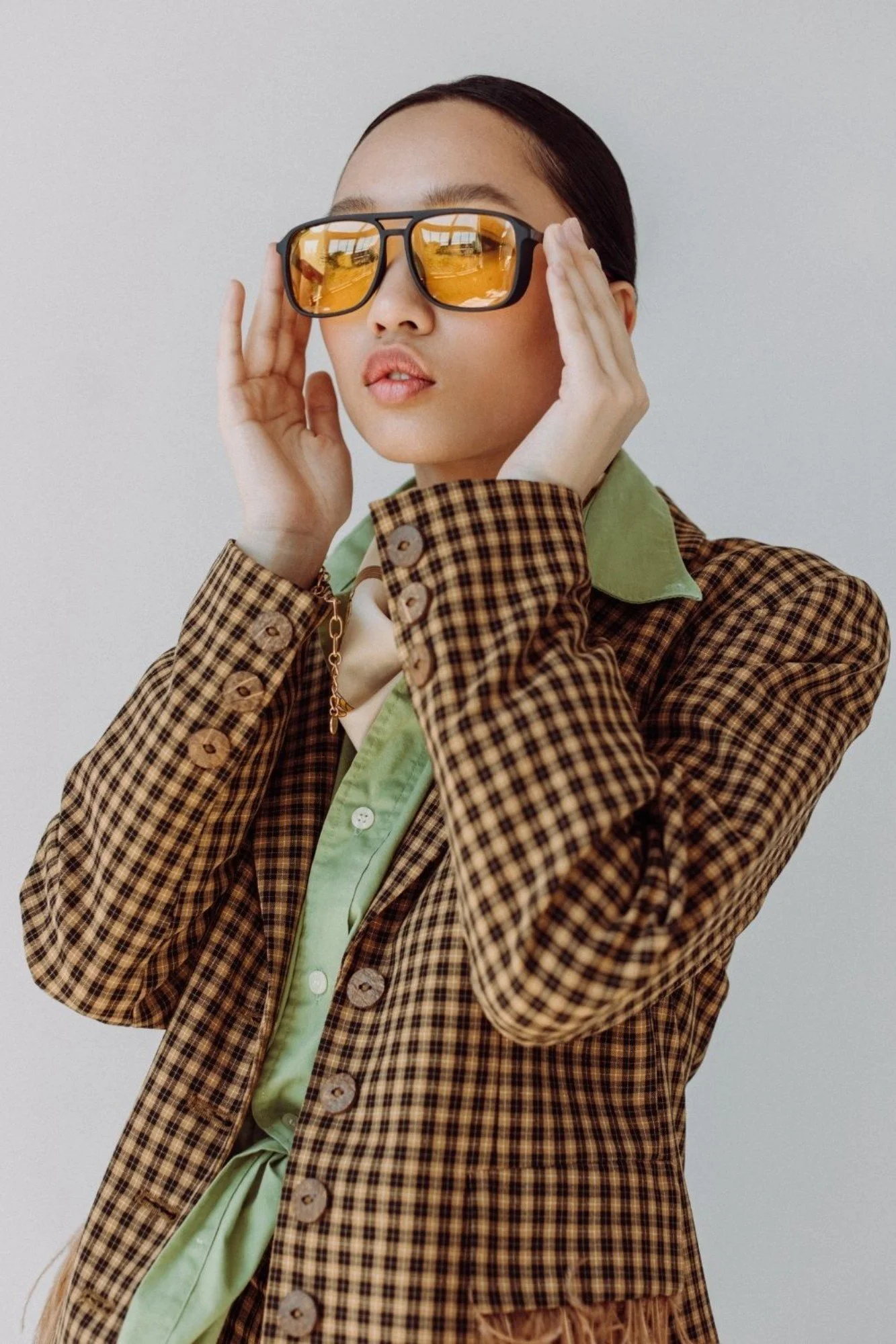A woman with dark hair wearing orange-tinted sunglasses, a plaid blazer, and a light green shirt, standing against a plain background.