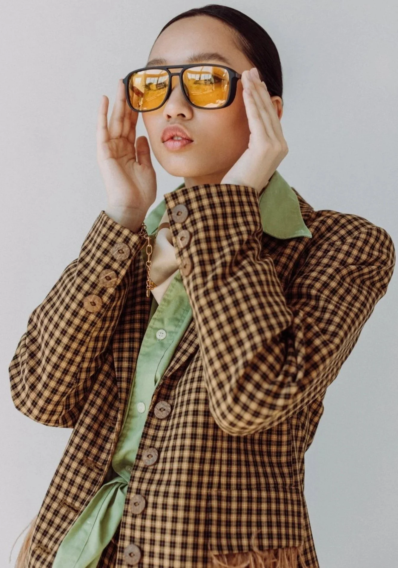 A woman wearing large sunglasses with yellow-tinted lenses, a green collared shirt, and a checked brown and black blazer.