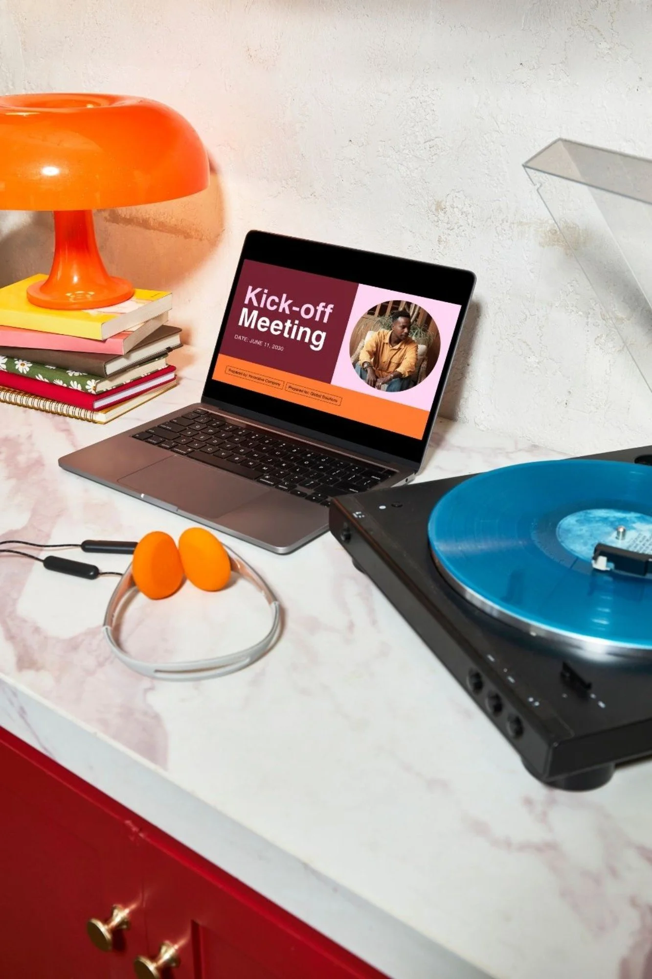 A workspace with a laptop displaying a 'Kick-off Meeting' presentation, a turntable with a blue vinyl record, orange headphones, a red and orange table lamp, a stack of colorful books, and a white marble countertop.