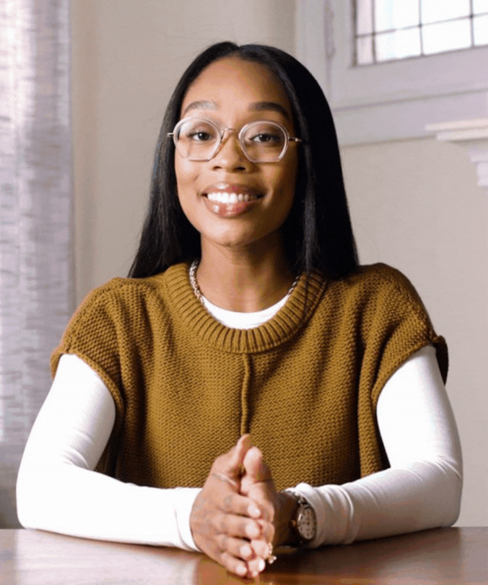 A young woman with long black hair, wearing glasses, a white long-sleeve shirt, and a brown knitted vest, smiling and sitting at a table indoors.