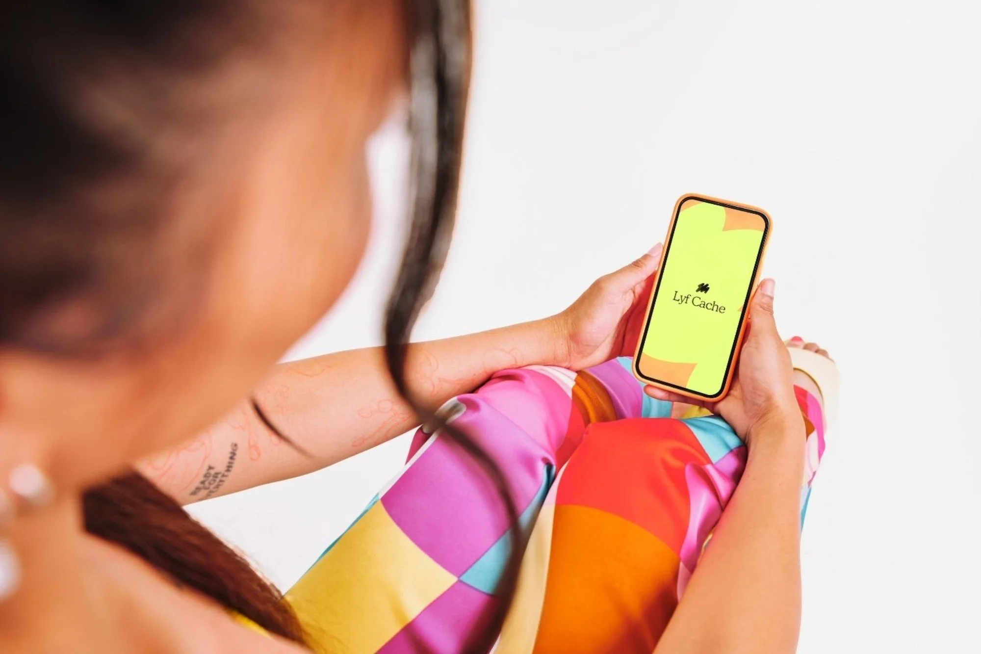 A woman sitting and holding a smartphone that displays the Lyft Cache app, wearing colorful patterned pants and a sleeveless top.