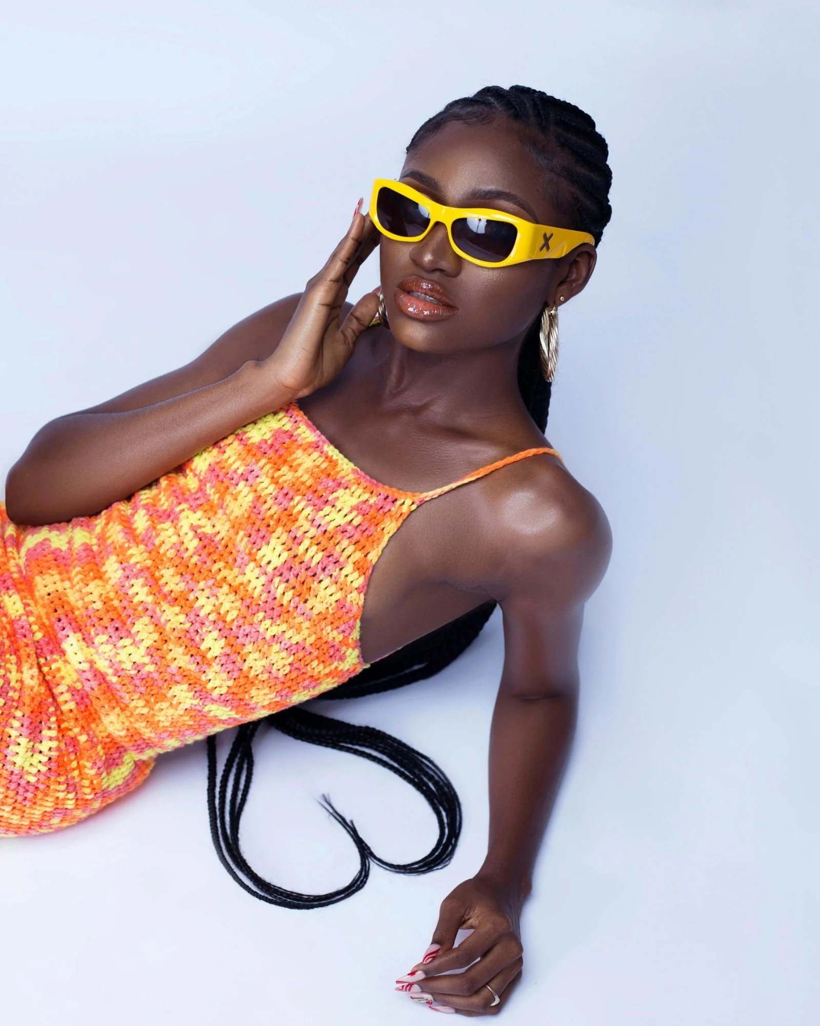 A woman wearing yellow sunglasses, a colorful knitted dress, and hoop earrings, lying on a white surface with her long braids visible.