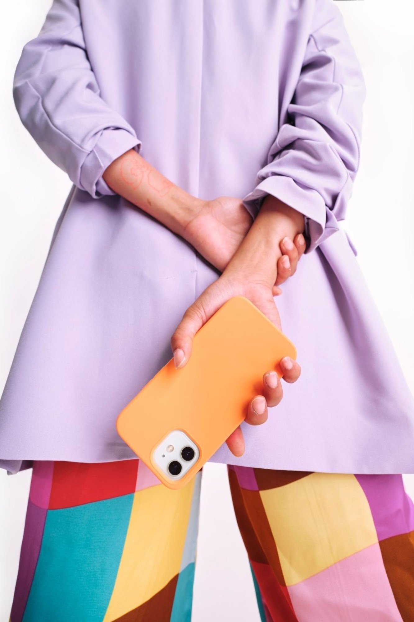 Person wearing a lavender coat and multicolored patchwork pants holding a yellow phone with a case behind their back.