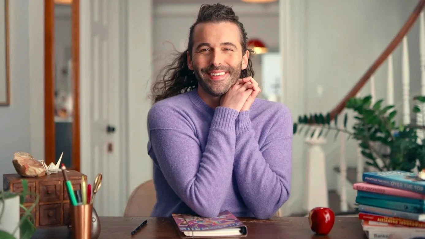A man with long hair, beard, and mustache, smiling and sitting at a desk with a purple sweater, with a closed notebook, pen, apple, books, and office supplies on the desk, in a well-lit room with plants and stairs in the background.