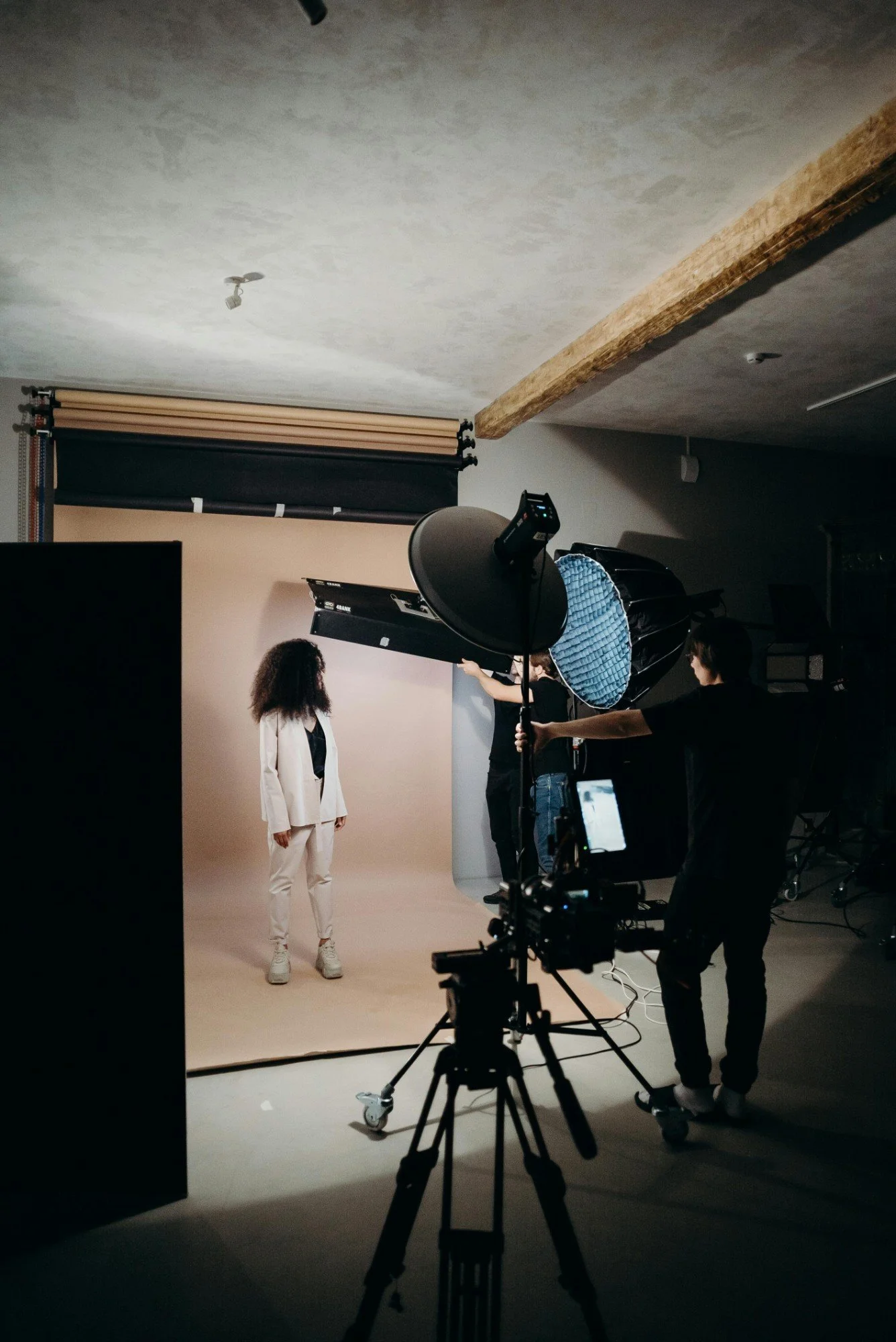 A photoshoot setup with a woman in a white suit and sneakers standing in front of a beige backdrop. Photographers and lighting equipment are positioned around her inside a studio.