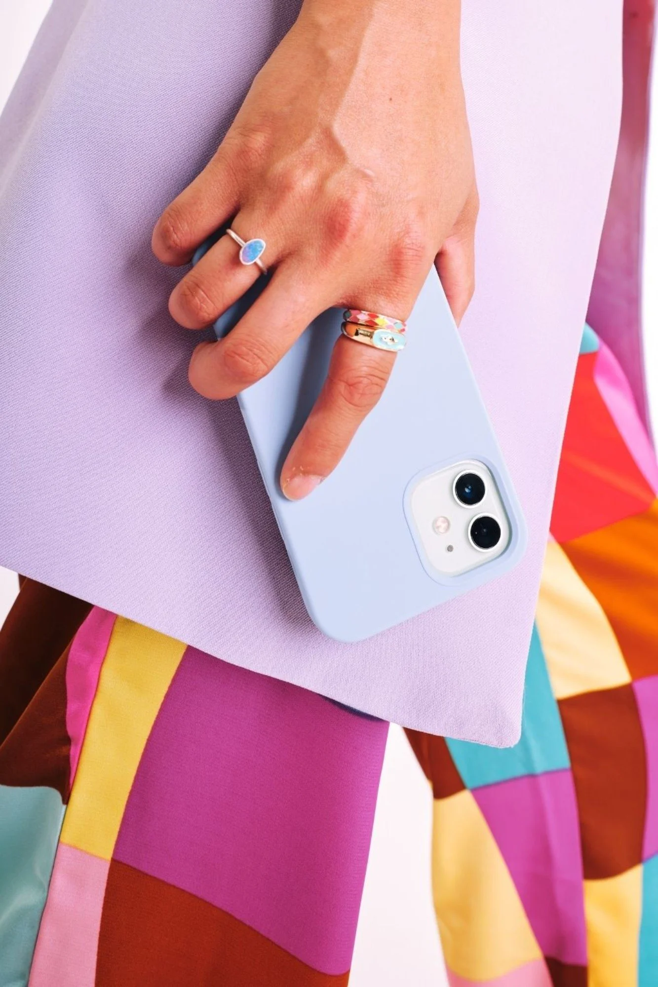 Close-up of a person's hand holding a light blue smartphone, wearing colorful rings, and standing against a pastel purple clutch and multicolored patterned clothing.