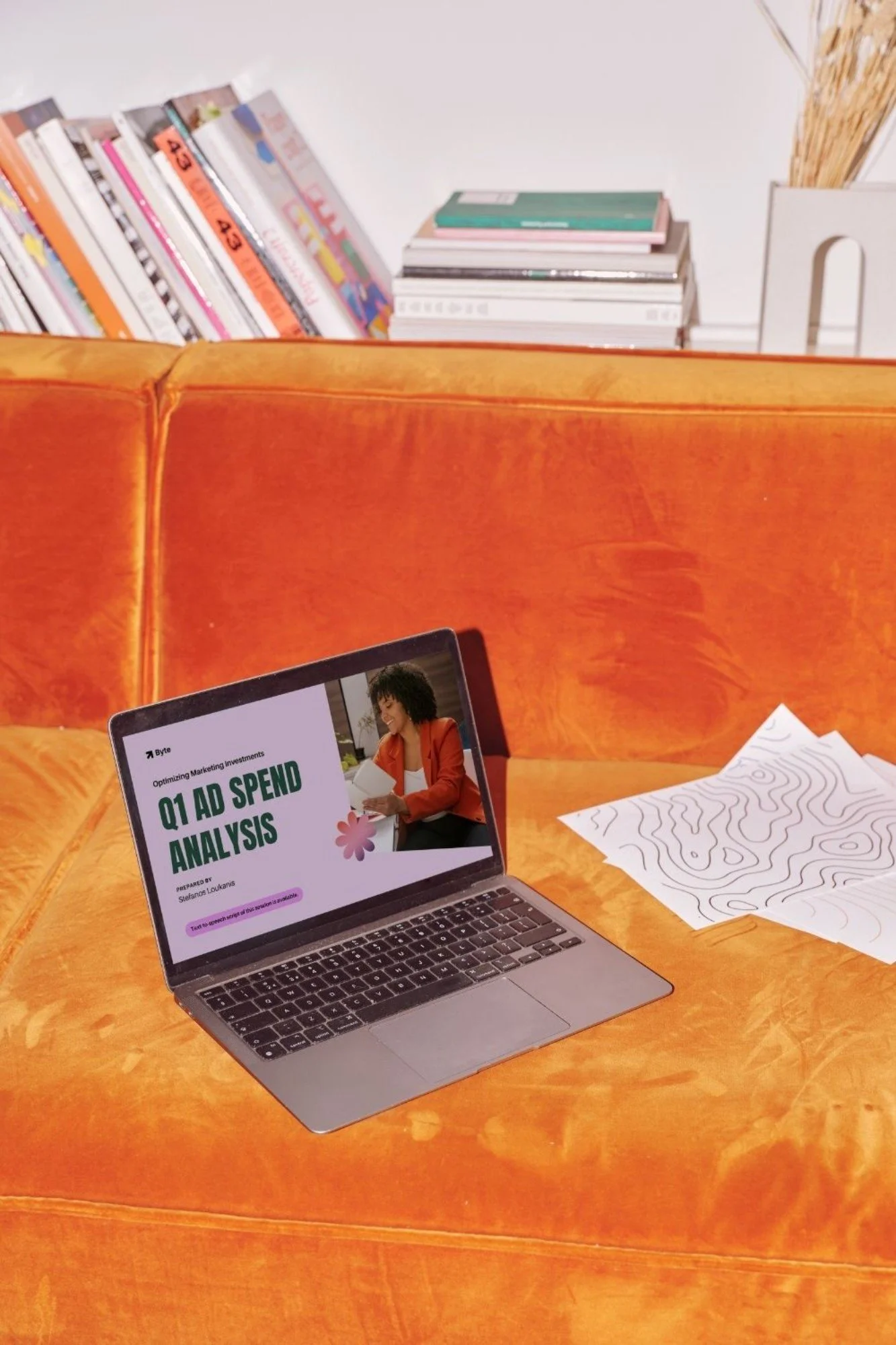 A laptop on a bright orange velvet couch displaying a presentation titled 'Q1 AD SPEND ANALYSIS' with a woman reading a paper on the slide. Papers with line drawings are on the couch to the right, and a stack of magazines or books is placed on the back of the couch with more books or magazines on a shelf in the background.