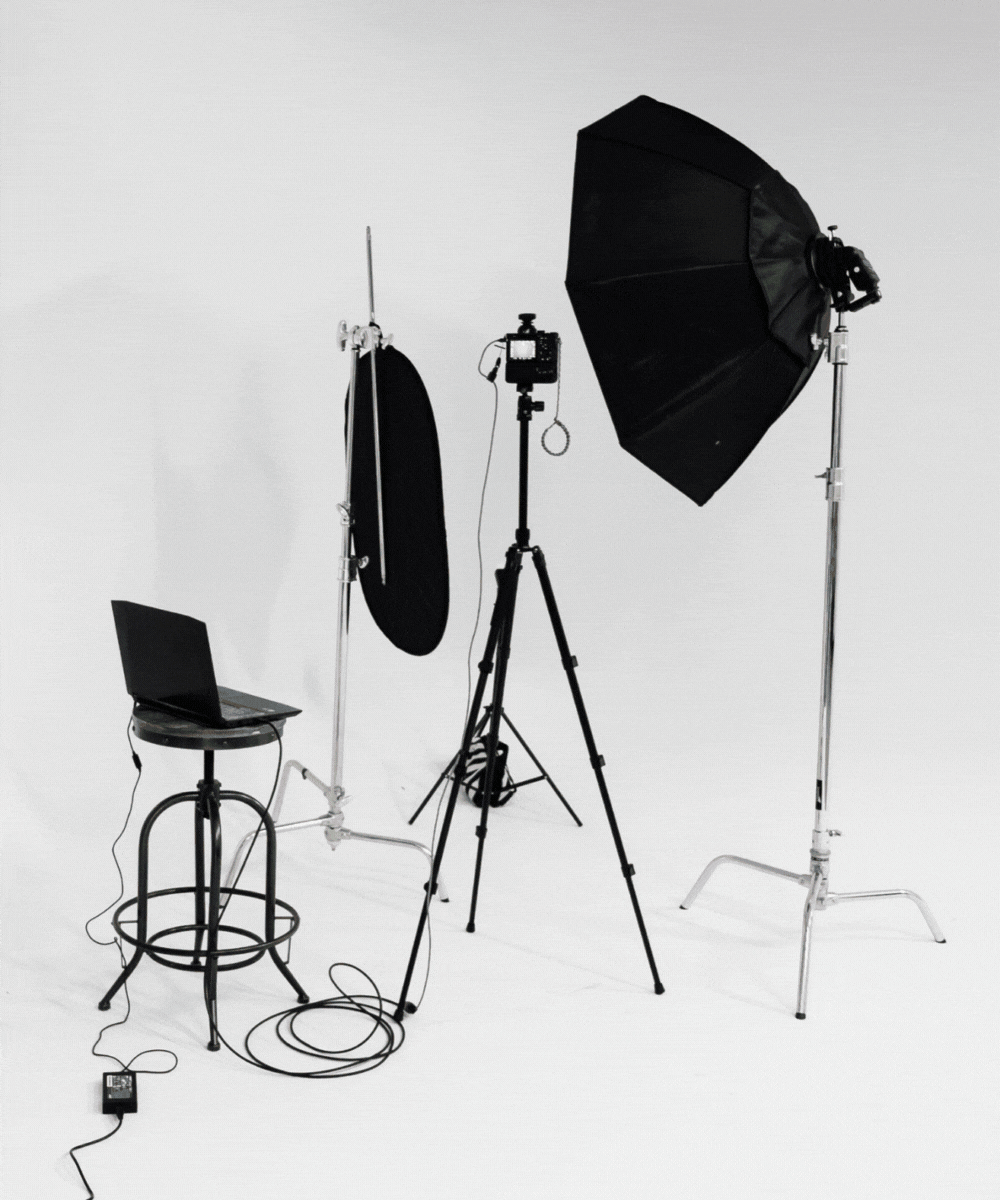 Photo of a photography studio setup with lighting equipment, a camera on a tripod, a softbox, reflector, laptop on a stool, and an extension cord on a white background.