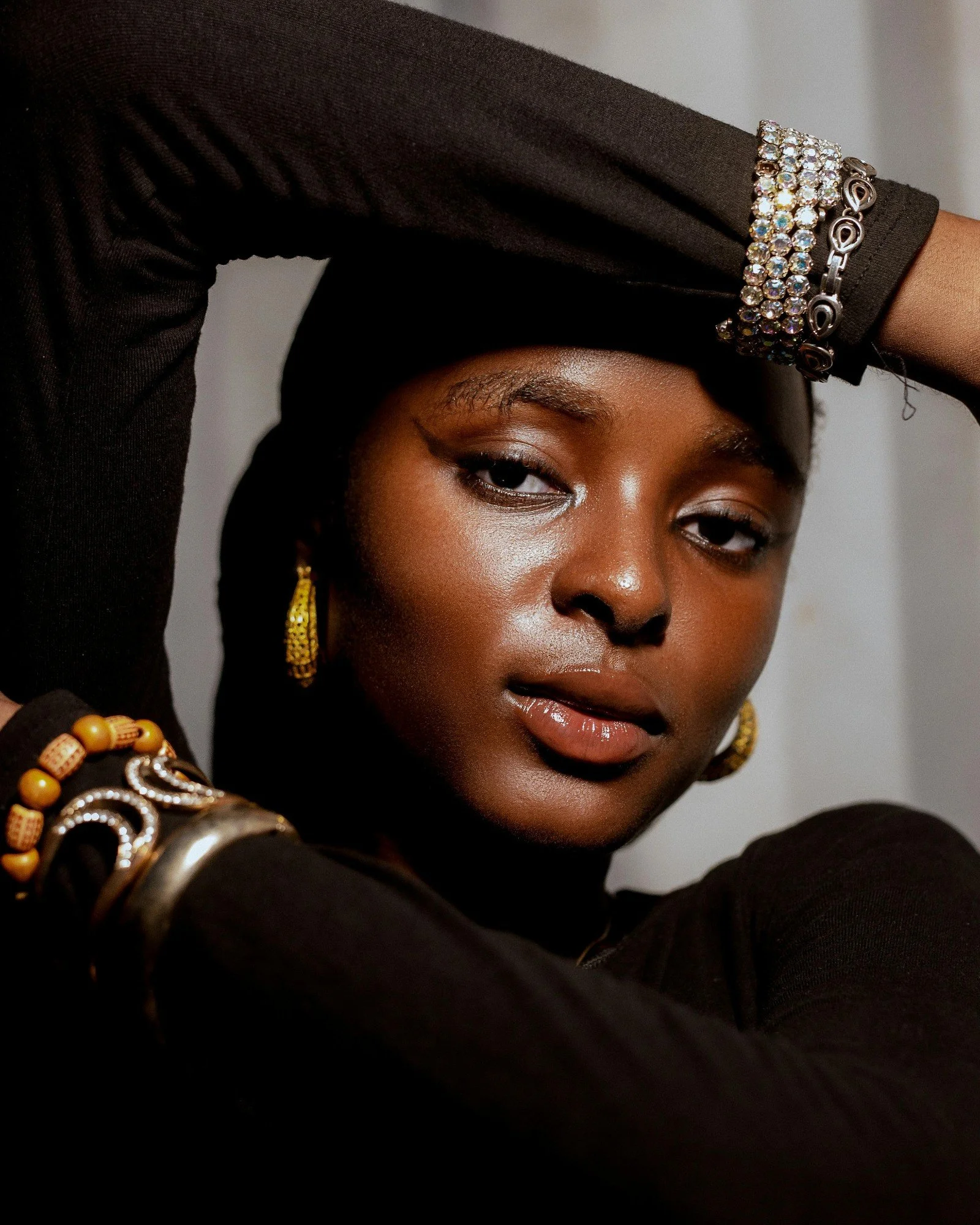 Close-up portrait of a woman with dark skin, wearing a black top and various jewelry including earrings and bracelets, with a serious and confident expression.