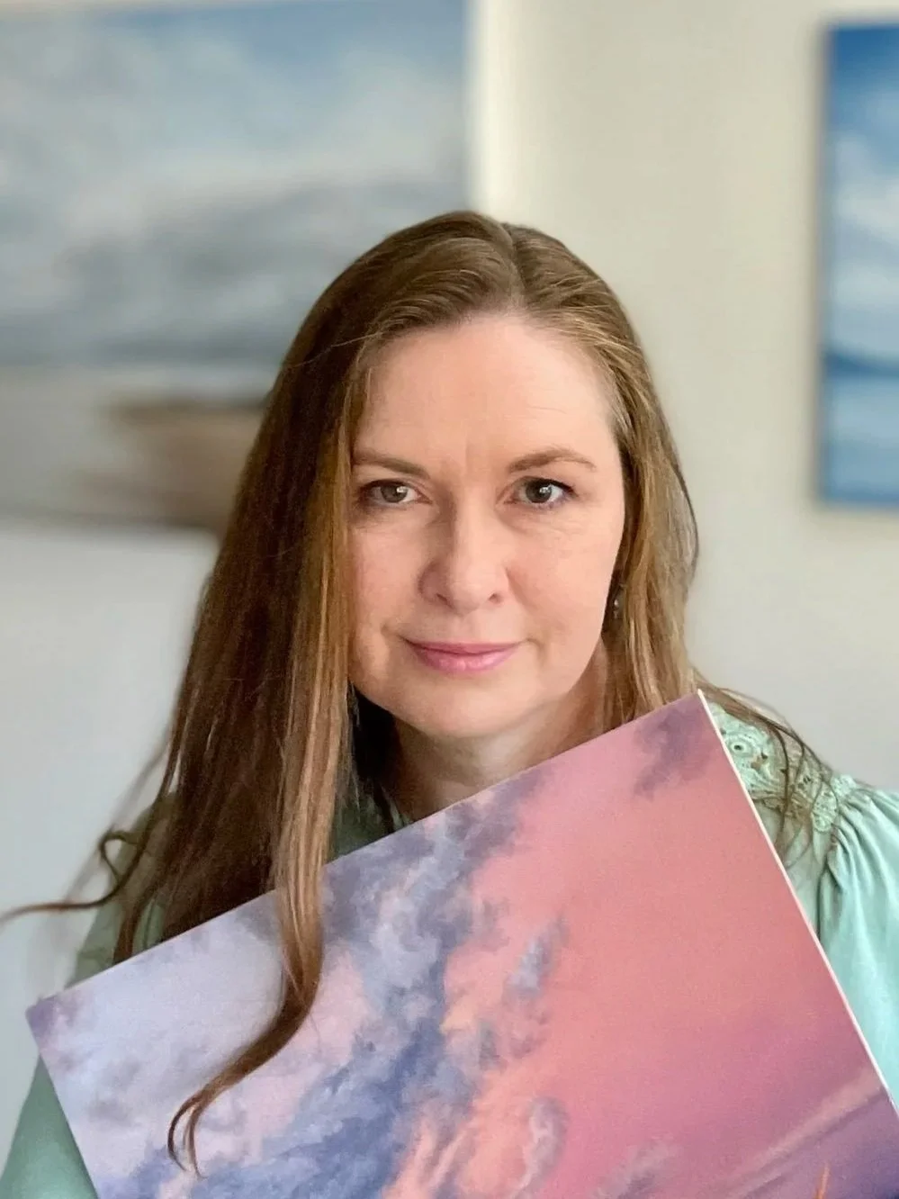 A woman with long light brown hair and light skin. She is holding a colorful item with pink and purple hues and looking at the camera. Blurred blue and gray artwork is in the background.