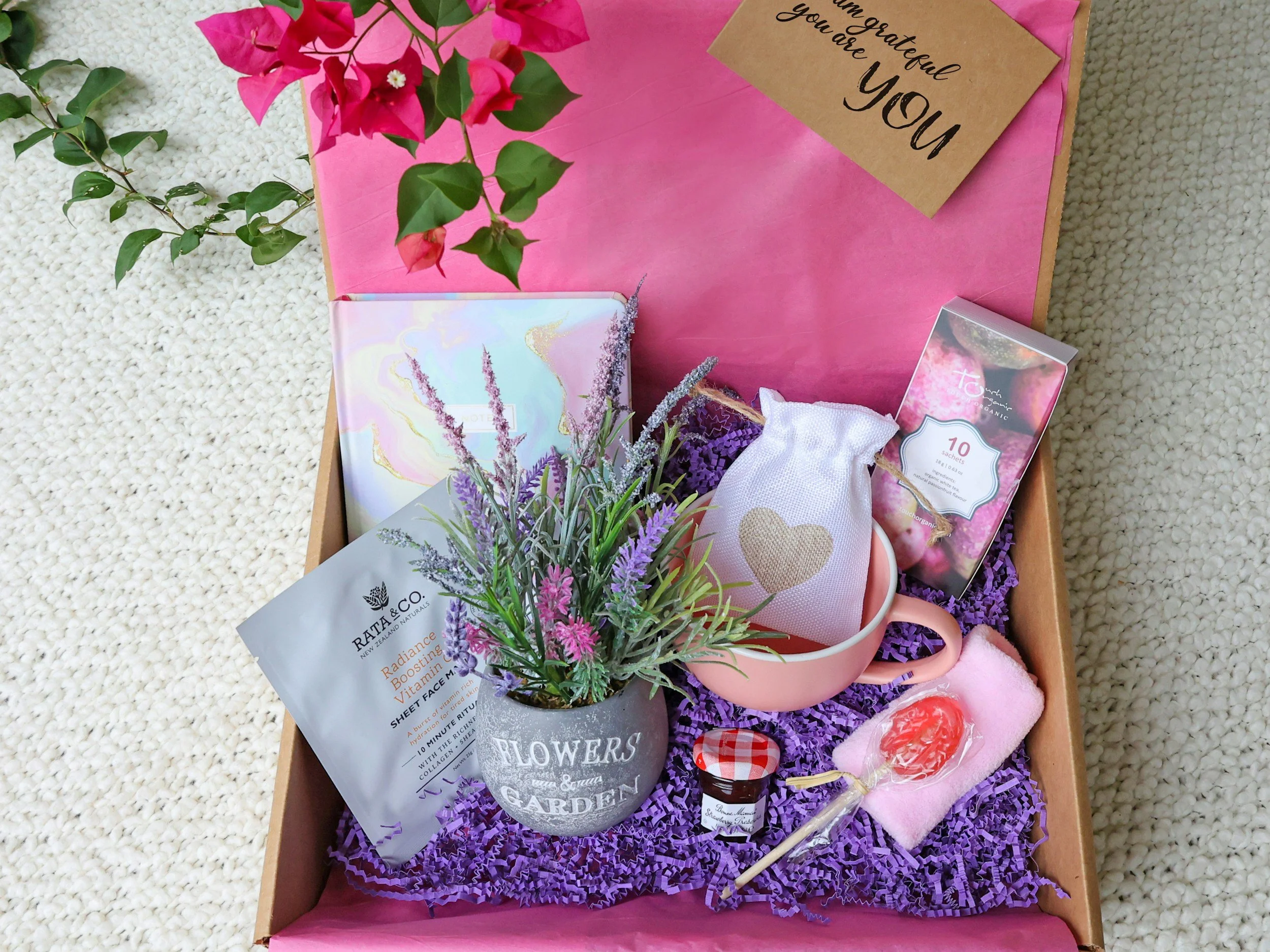 A gift box with various items including a pink mug, a small bag with a heart, a box of tea, a bundle of lavender, a sheet face mask, a jar of jam, a pink lollipop, and an arrangement of pink flowers and greenery. Inside the box is purple shredded paper and a pink paper with a thank you message.